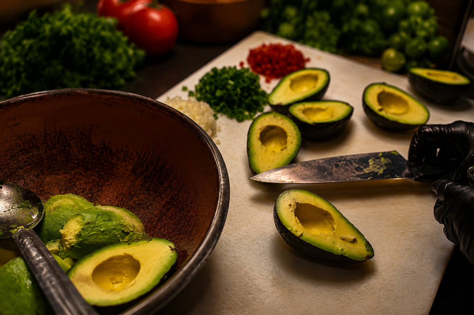Halved avocados on a cutting board with a knife, green vegetables and tomatoes in the background, preparing for food preparation.