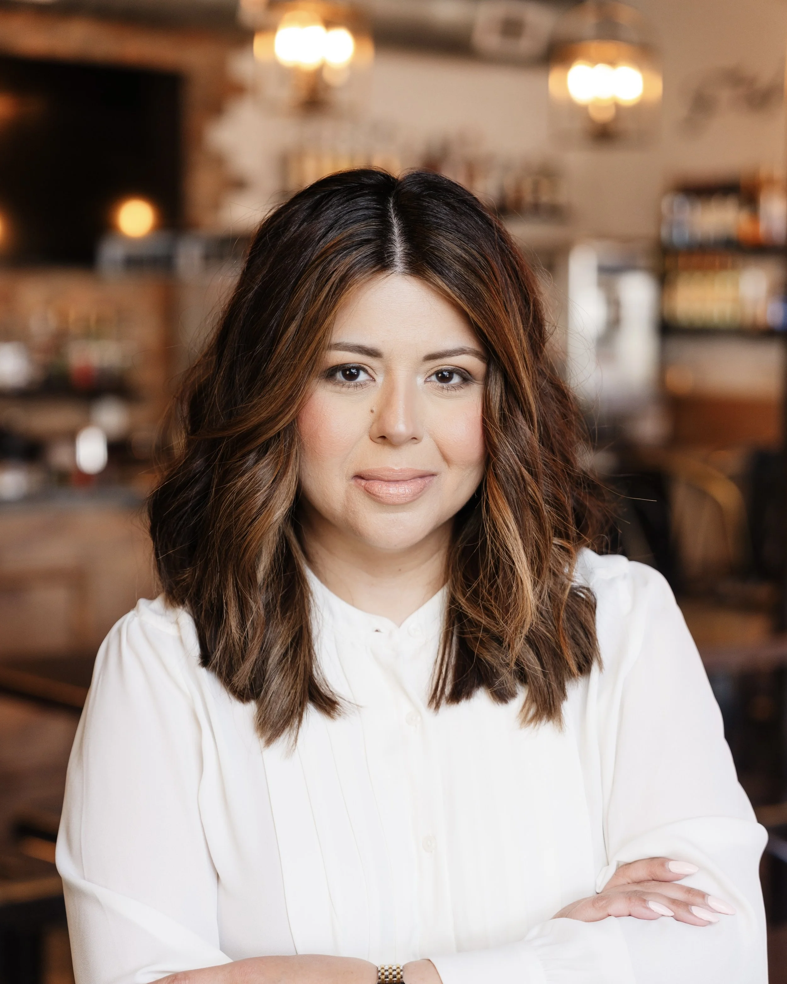 Close-up of a woman with long, wavy brown hair with highlights, wearing a white blouse, standing with arms crossed in a warmly lit room with blurred background of walls, shelves, and hanging lights.