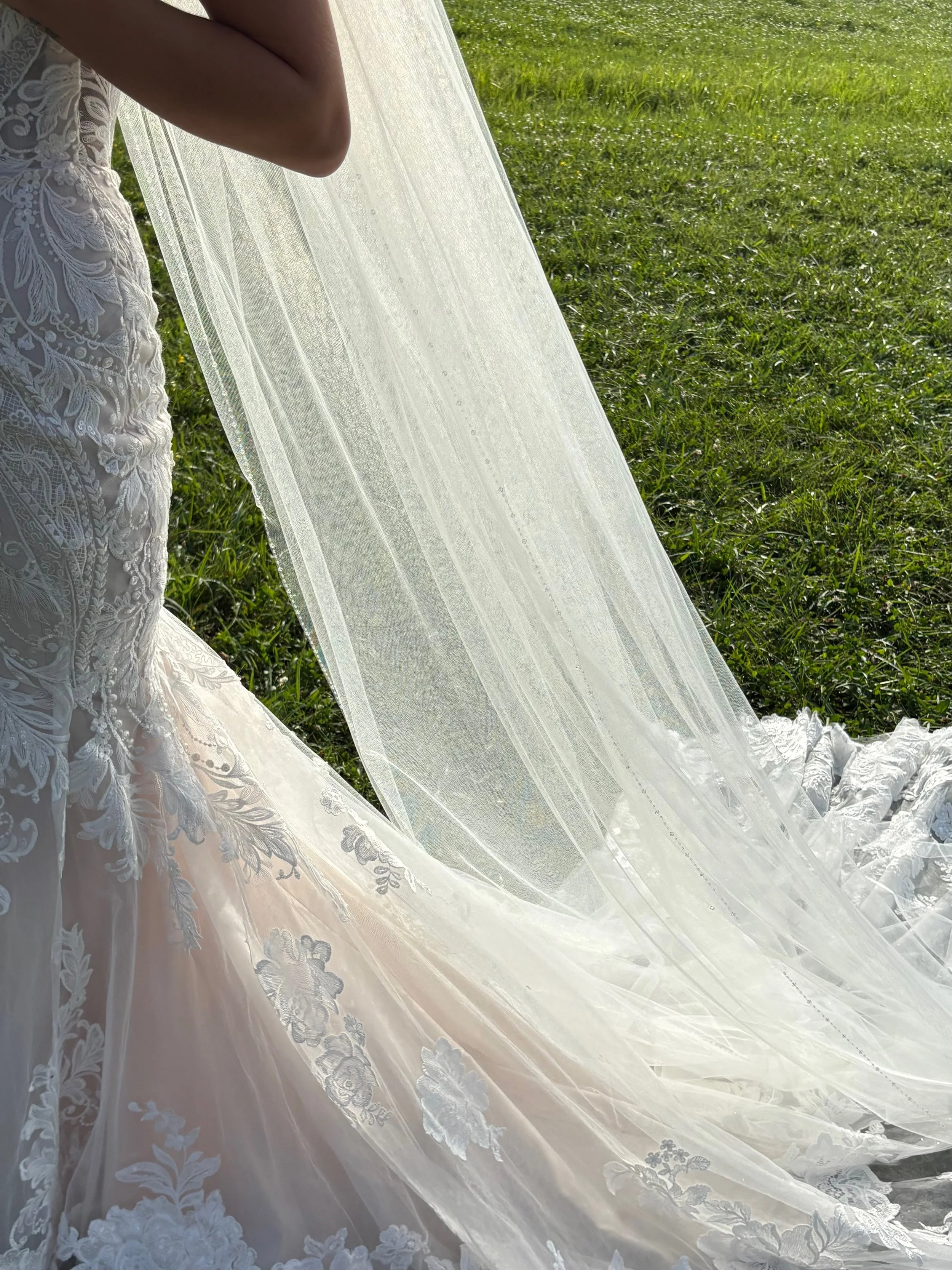 Close-up of a white wedding dress with intricate lace embroidery, lying on the grass with a sheer veil.