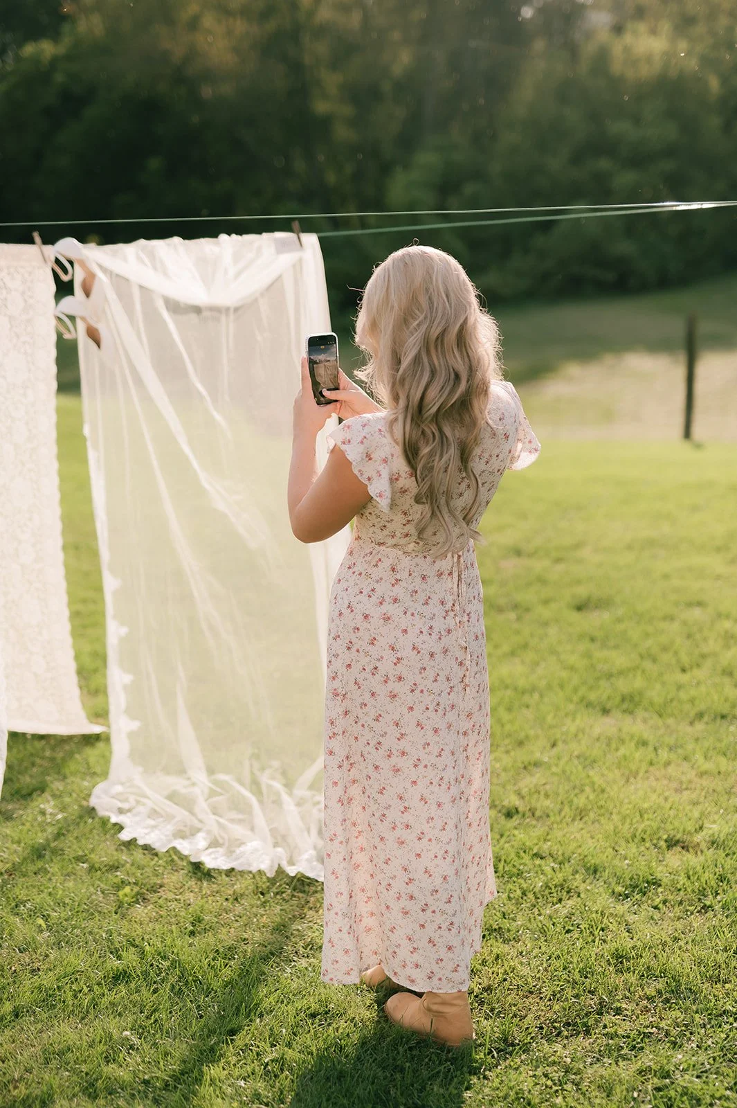 A woman with long, wavy blonde hair taking a photo with her phone while standing on a grassy field. She is wearing a long, floral-patterned dress and tan boots. Behind her, a line of white lace and sheer fabric is hanging on a clothesline outdoors.