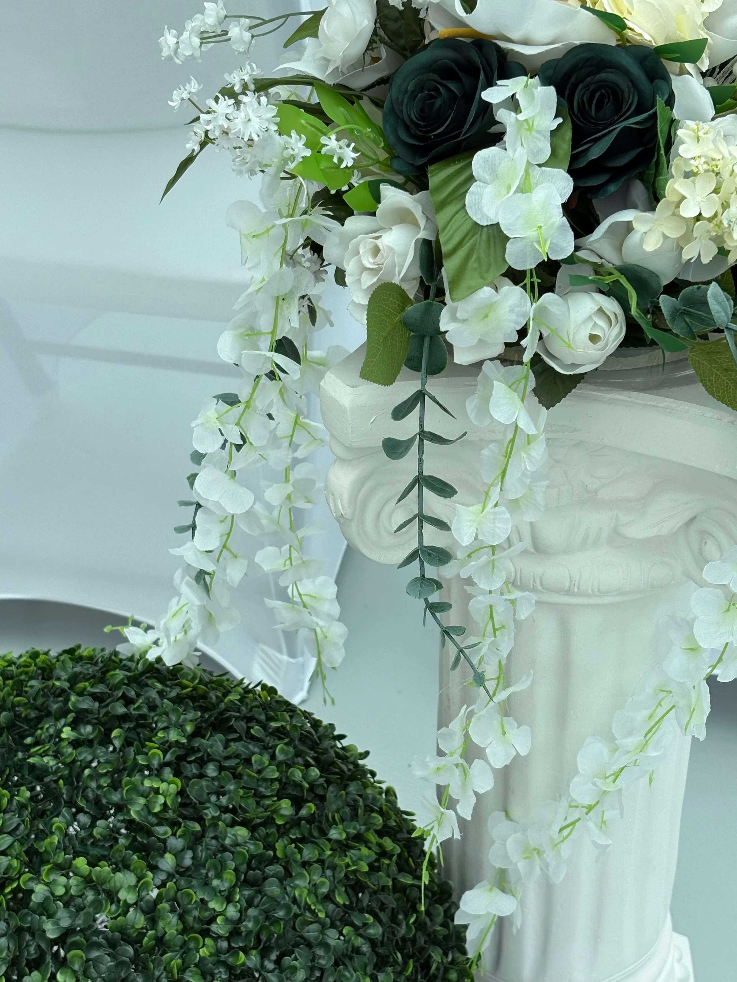A decorative floral arrangement featuring white and dark green flowers, green leaves, and white cascading vines in a white vase, with a small green bush in front.