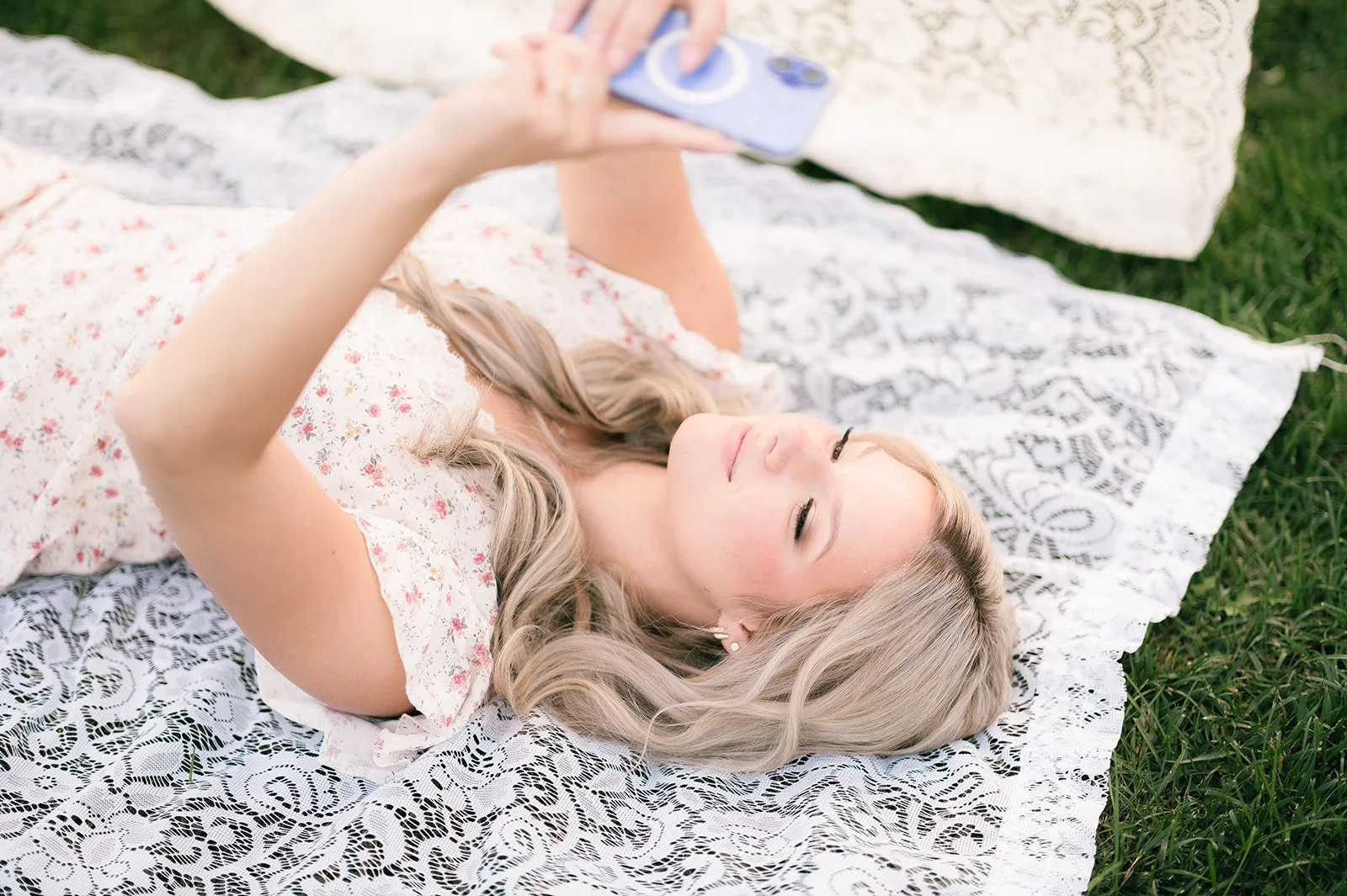 A young woman with blonde hair lying on a lace blanket on grass, looking at her phone with a relaxed expression.