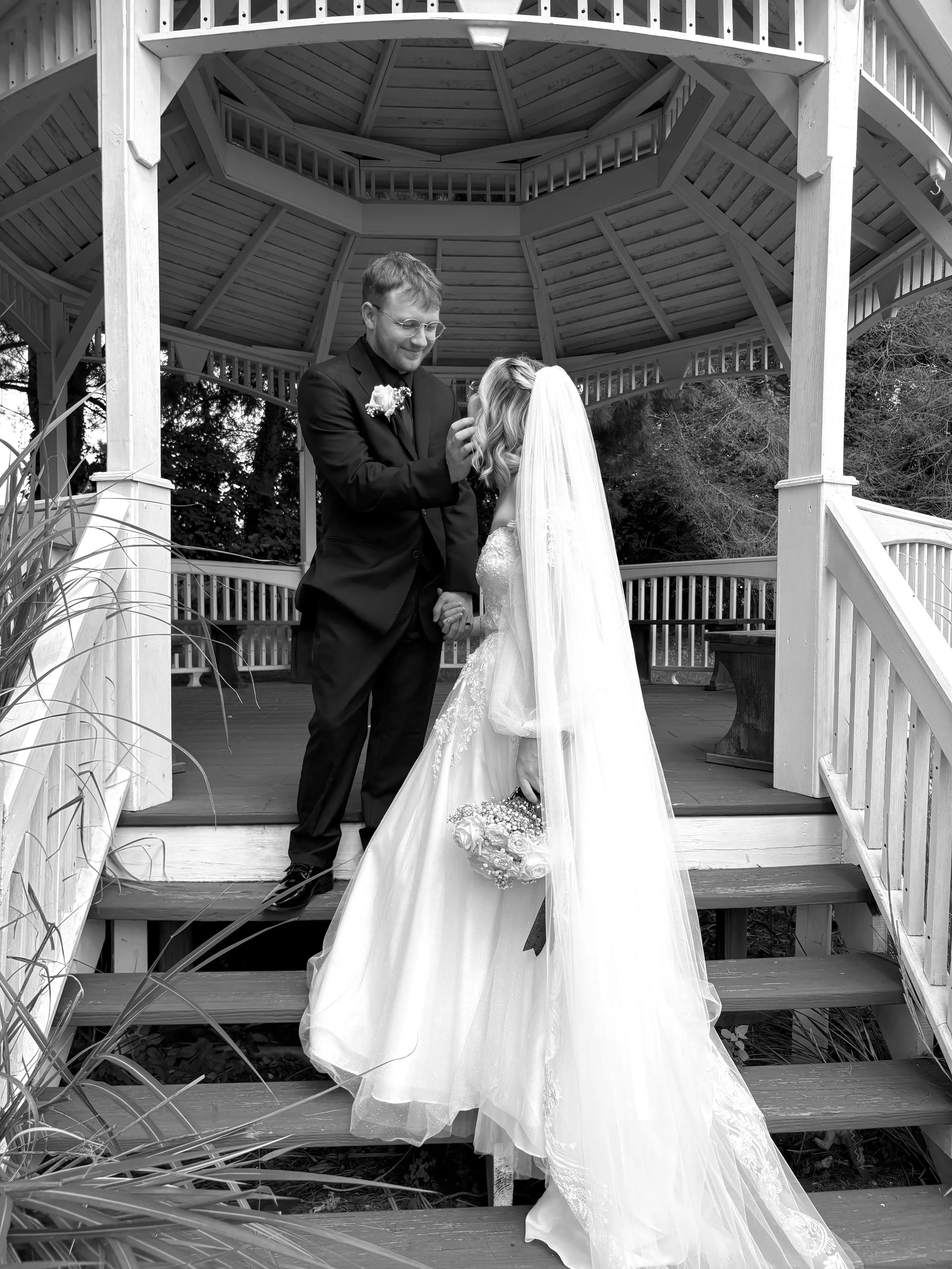 Black and white photograph of a bride and groom standing on a wooden gazebo, holding hands and facing each other. The groom is placing a ring on the bride's finger. The bride is wearing a wedding gown with a long veil and holding a bouquet of flowers. The gazebo features a multi-level roof and railings.