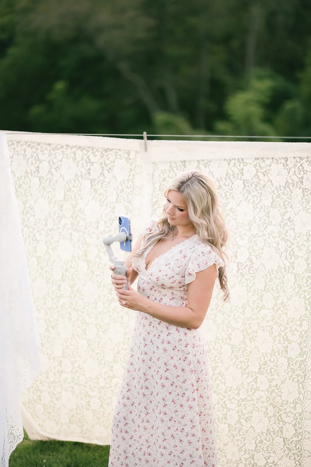 Woman in a floral dress taking a selfie with a phone gimbal outdoors with a lace curtain backdrop and green trees in the background.