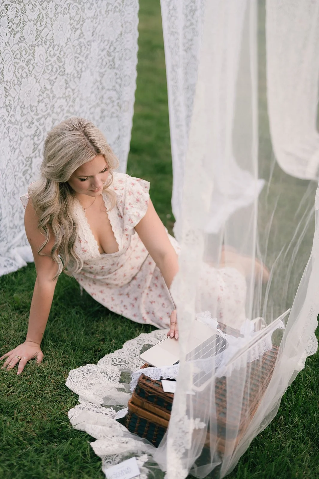 A woman with long blonde hair wearing a floral dress sitting on grass near lace curtains and a picnic basket with a laptop on a lace blanket.