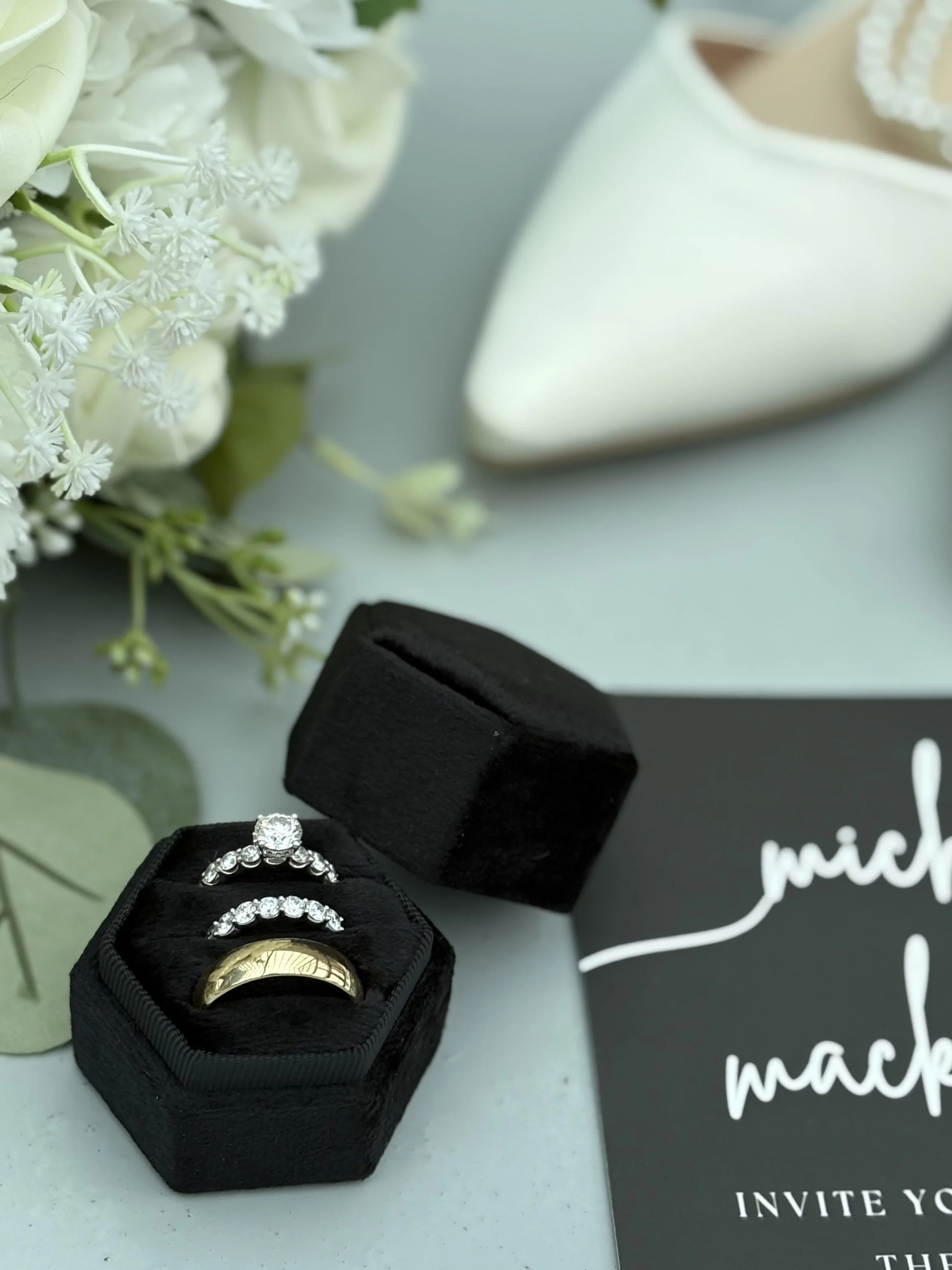 A black ring box holding a gold wedding band with an engraved design, a diamond engagement ring, and a diamond wedding band, with a bouquet of white flowers in the background, a black card with white text partially visible.