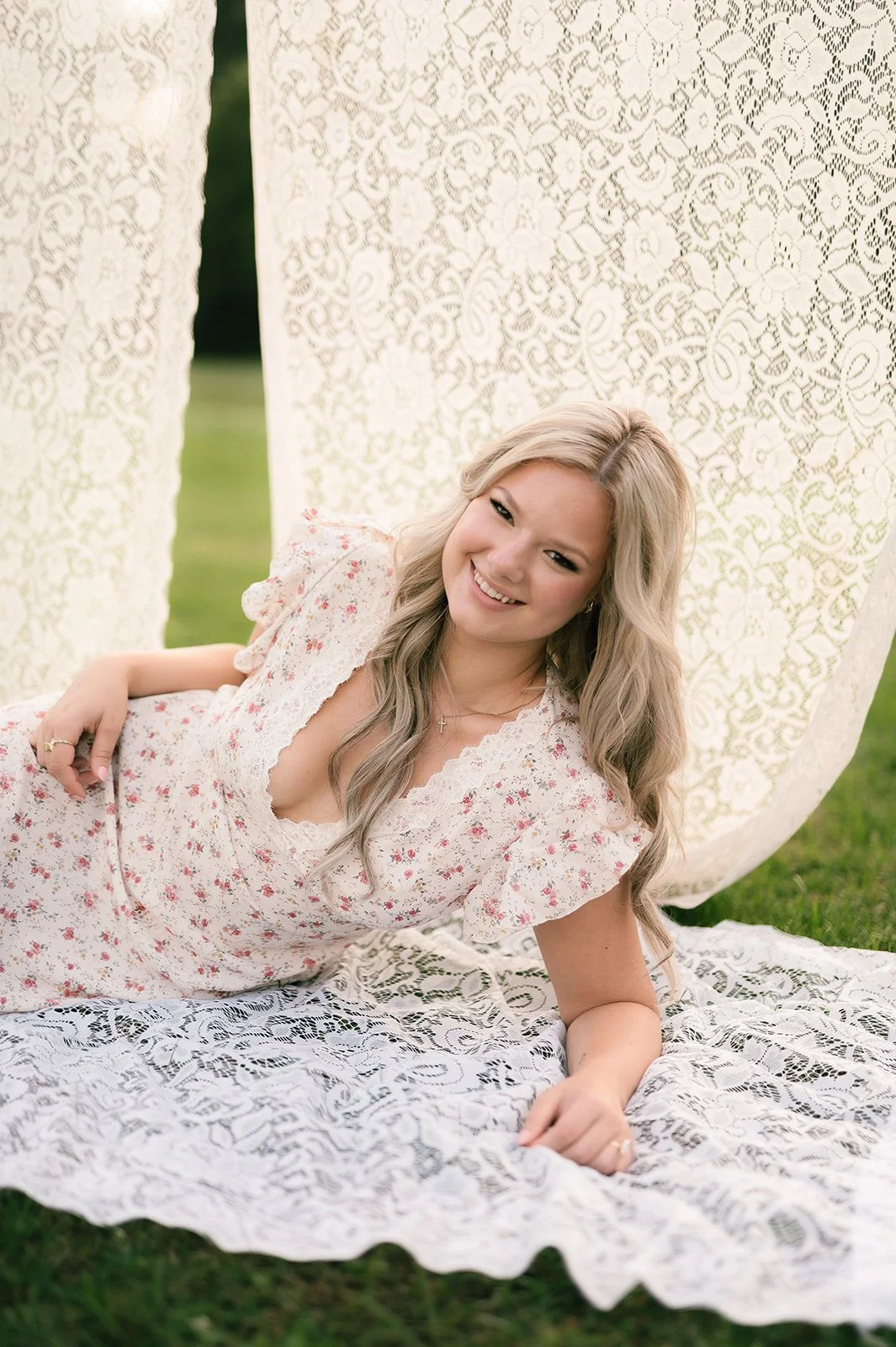 A woman with long wavy blonde hair lying on her side on a lace blanket outdoors, smiling at the camera with a lace backdrop behind her.
