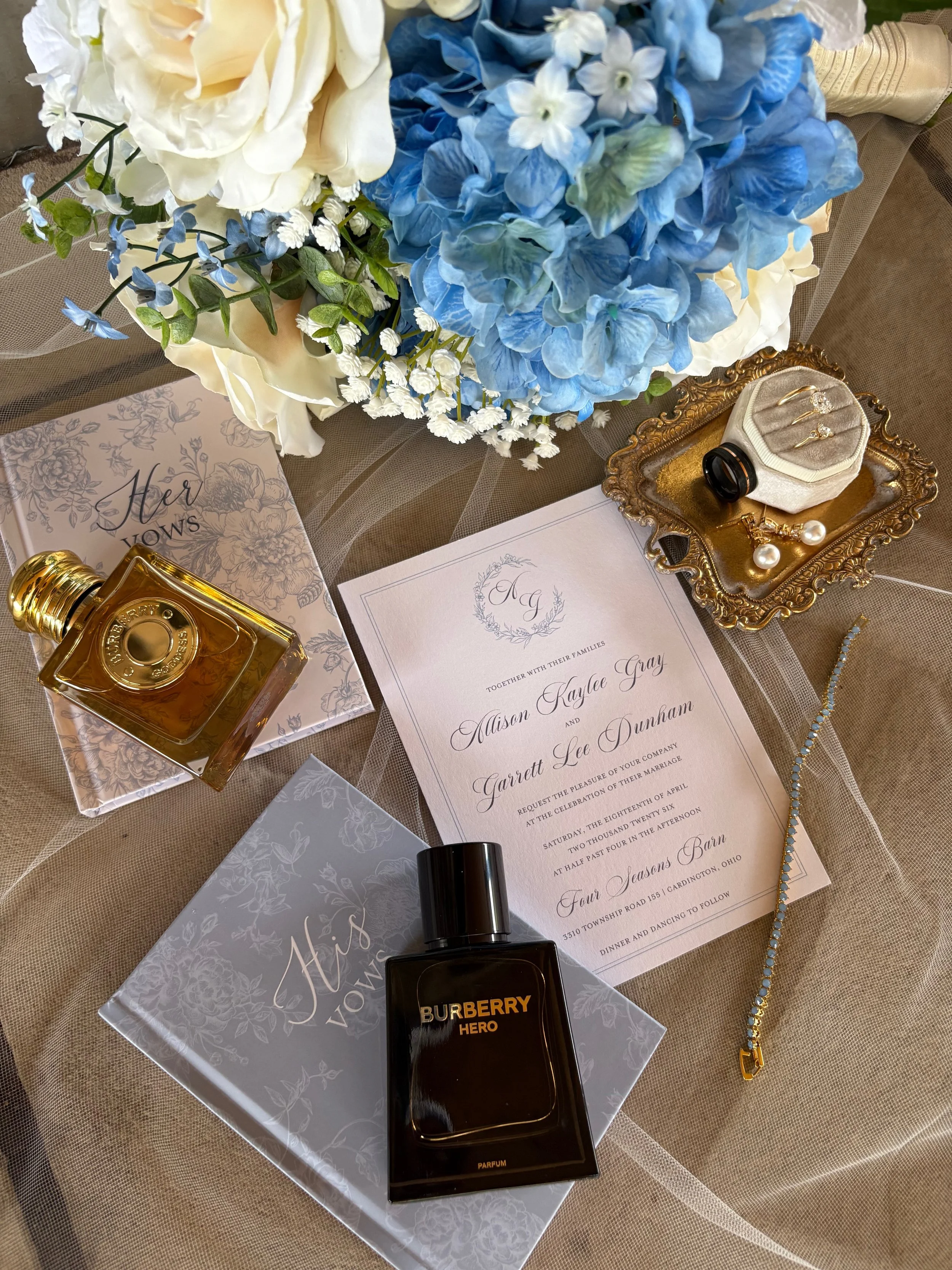 Wedding invitation surrounded by blue and white flowers, jewelry, perfume, and a ring box on a table.
