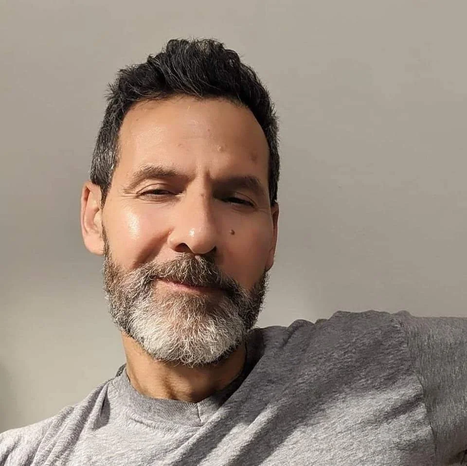 Close-up of a middle-aged man with salt-and-pepper hair and beard, smiling slightly with eyes partly closed, wearing a gray shirt and sitting against a plain wall.