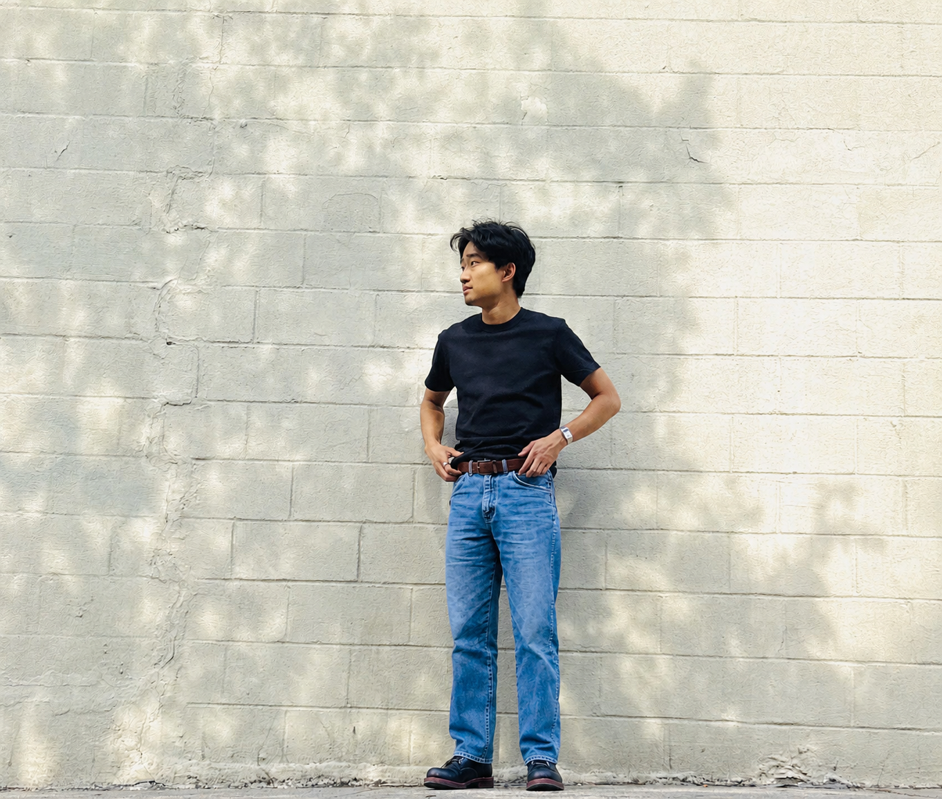 A young man standing against a beige brick wall, wearing a black t-shirt, blue jeans, a brown belt, and black shoes, looking to his left with his hands on his hips.