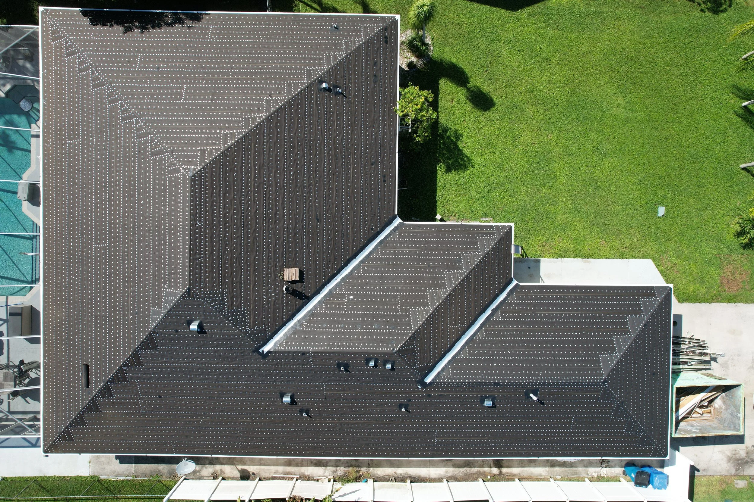 Aerial view of a residential building with a dark-colored roof covered in white solar panels, surrounded by a green lawn with trees and a swimming pool area on the left.