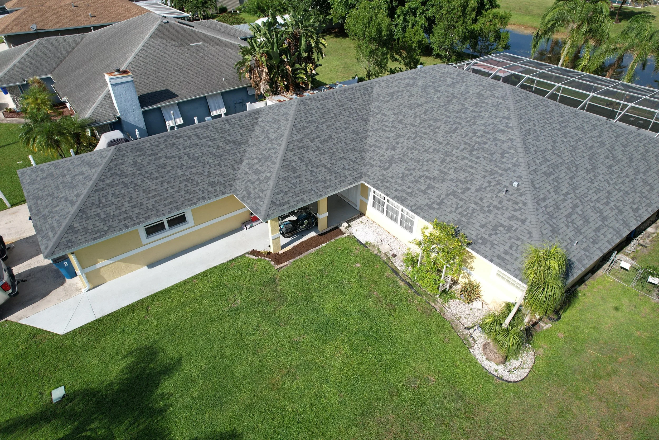 An aerial view of a house with a gray shingle roof, a backyard with green grass, trees, a pond, and a screened swimming pool.