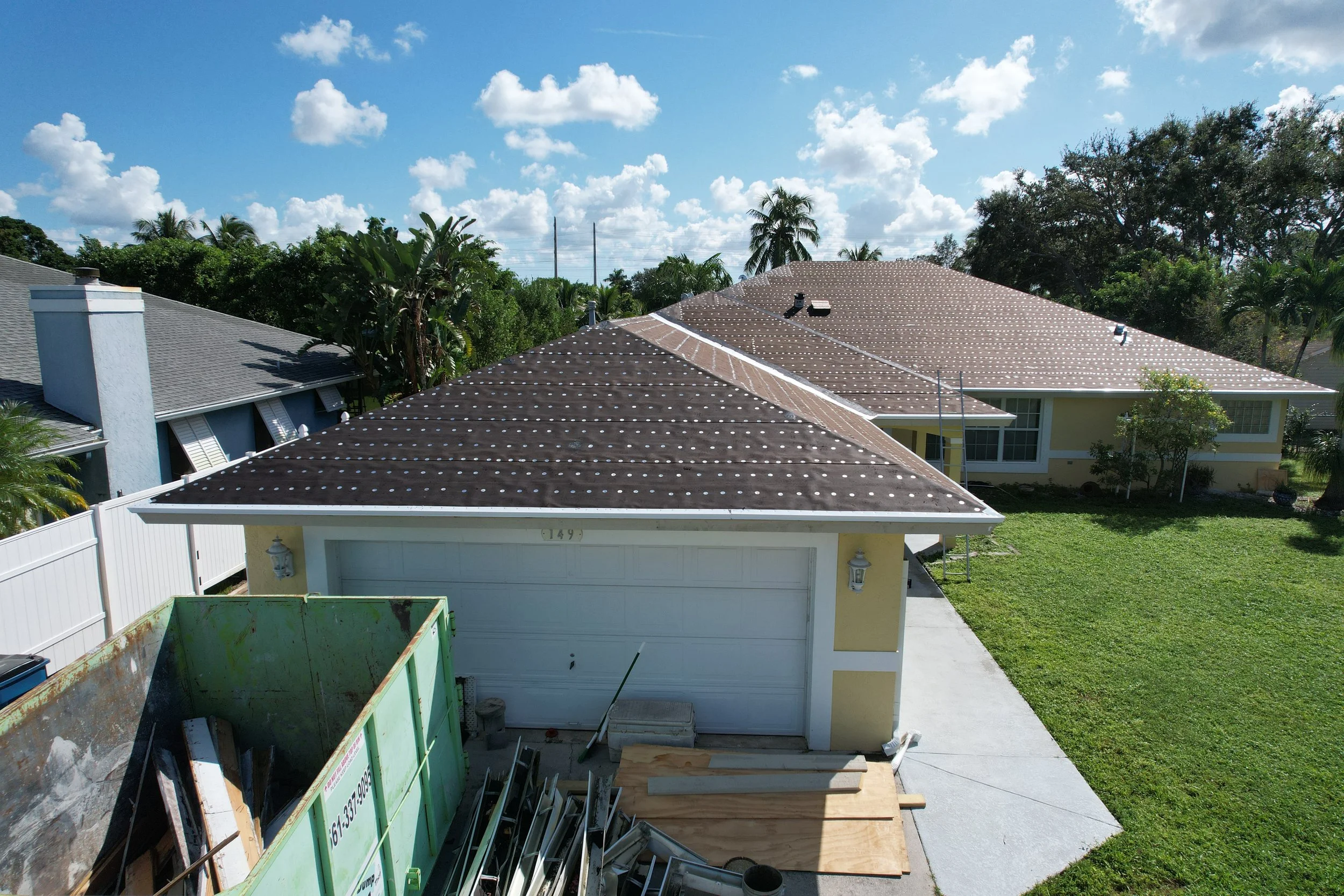View of rooftops of two houses with a blue sky and clouds. One house has a light yellow exterior, a white garage door, and a small window. The other house has a blue exterior. There are lush green trees and bushes surrounding the houses. A construction dumpster is in the foreground.
