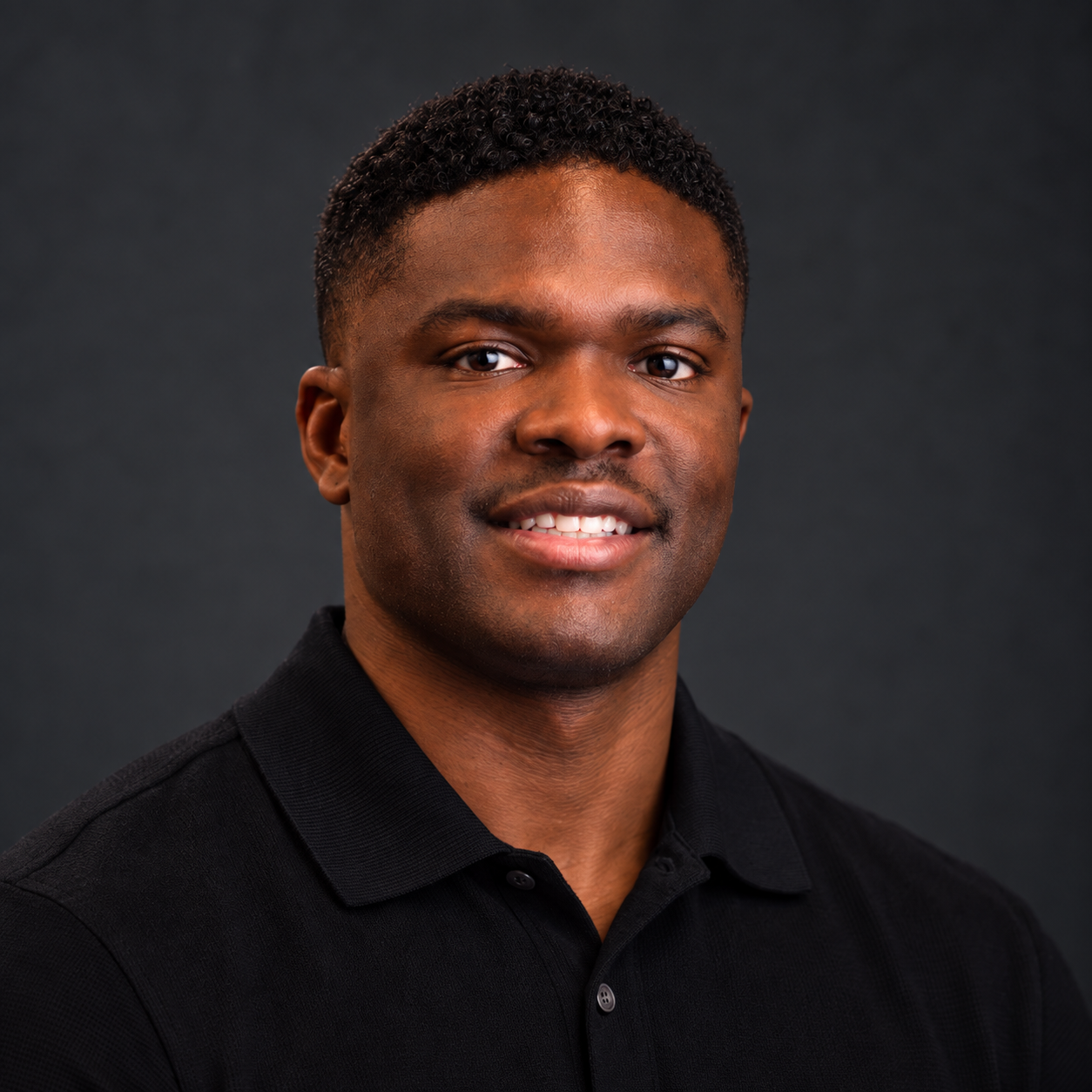 A young African American man with short, curly hair wearing a black collared shirt, smiling slightly, against a dark gray background.