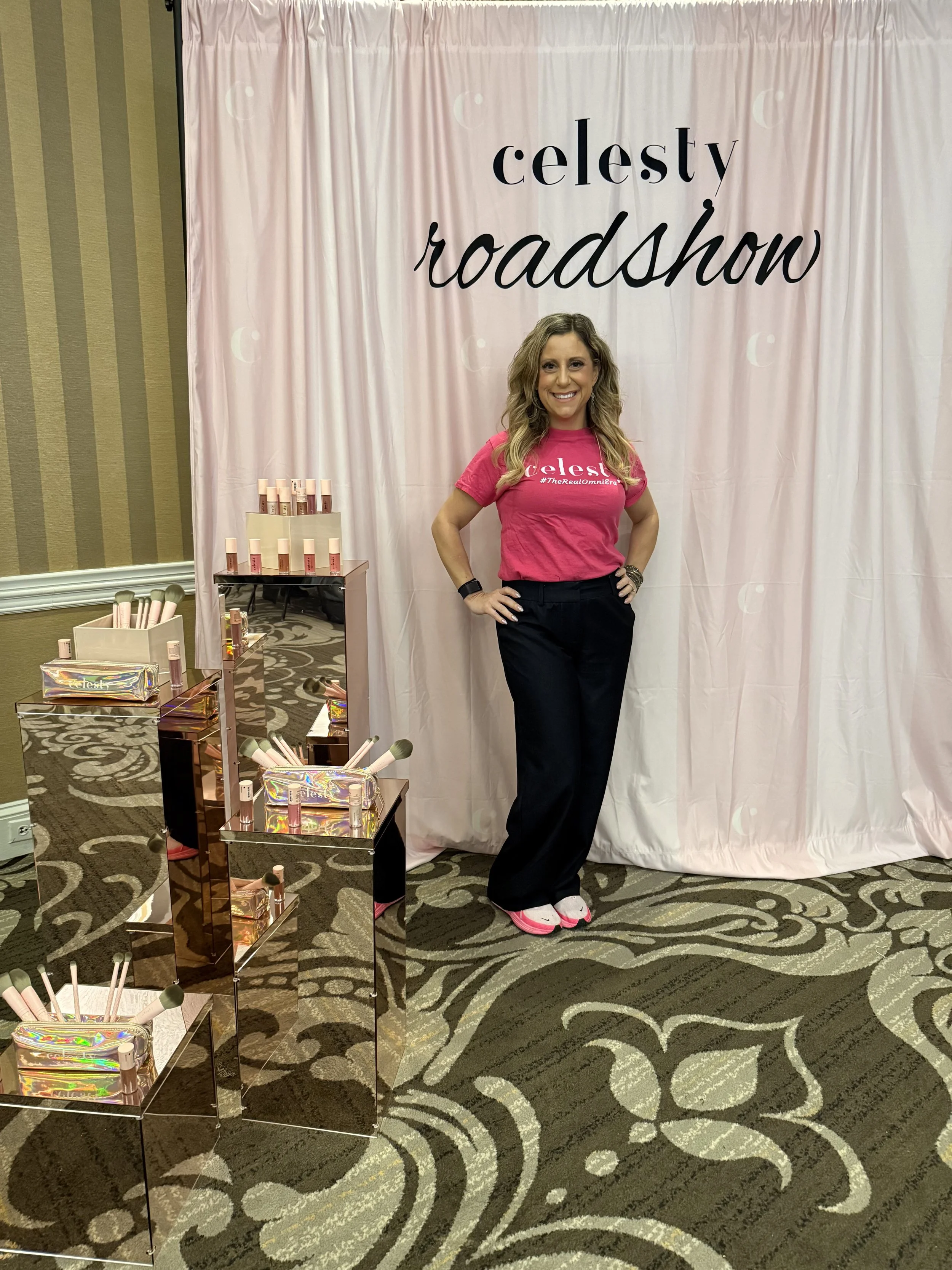 A woman standing in front of a white curtain with the text 'celestv roadshow' on it, smiling and wearing a pink t-shirt, black pants, pink shoes, and bracelets, with makeup and long wavy hair, at an event with display tables of skincare or makeup products.
