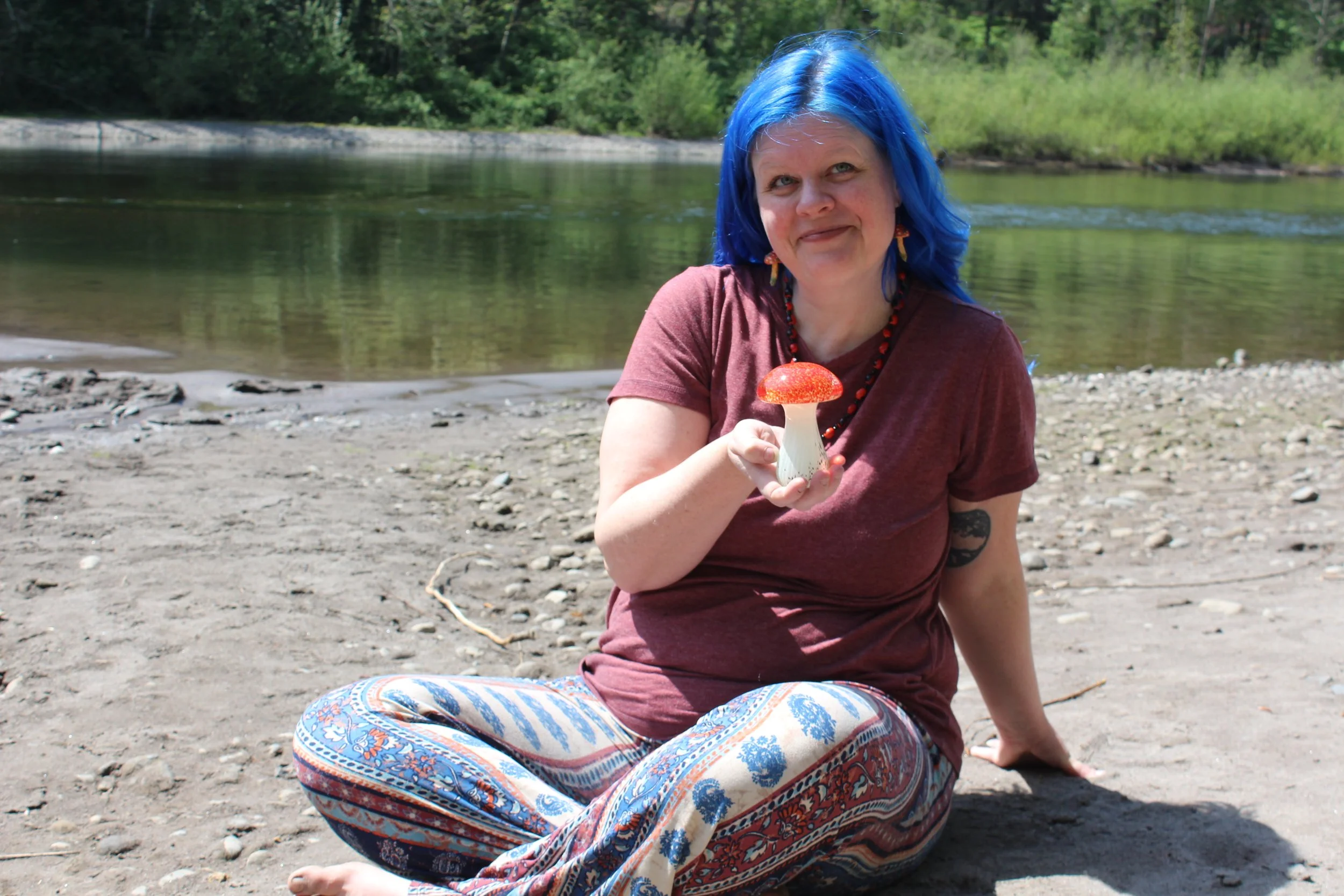 Woman with blue hair sitting on a rocky beach holding a red mushroom sculpture with a white stem, surrounded by trees and water.