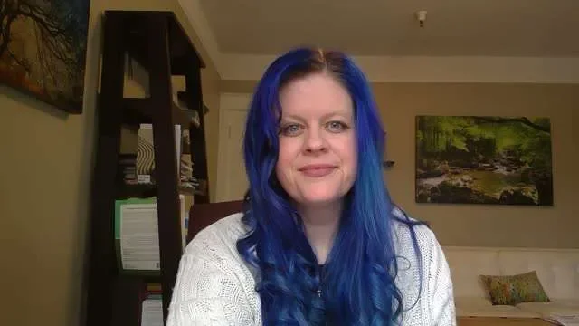 Woman with long, wavy, blue hair, smiling, sitting in a room with bookshelves and landscape pictures on the wall.