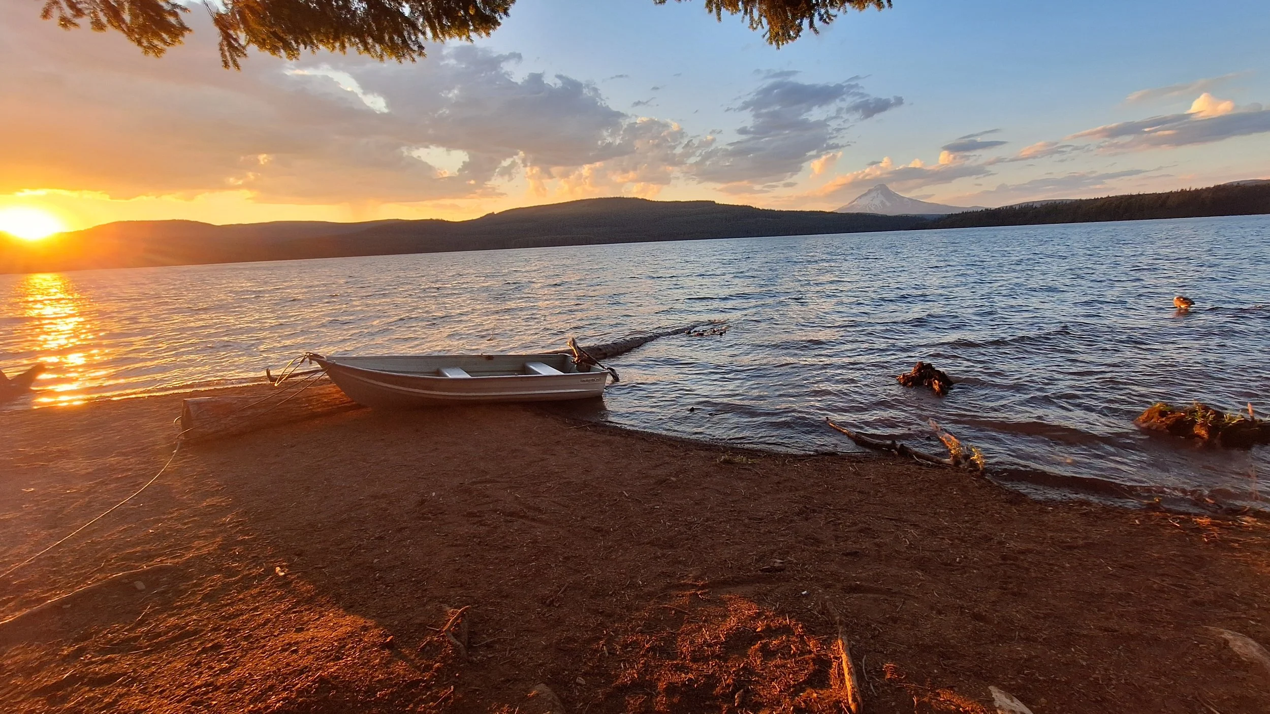 A boat resting on the sandy shore of a lake at sunset with mountains and a snow-capped peak in the background, and a sky filled with clouds.