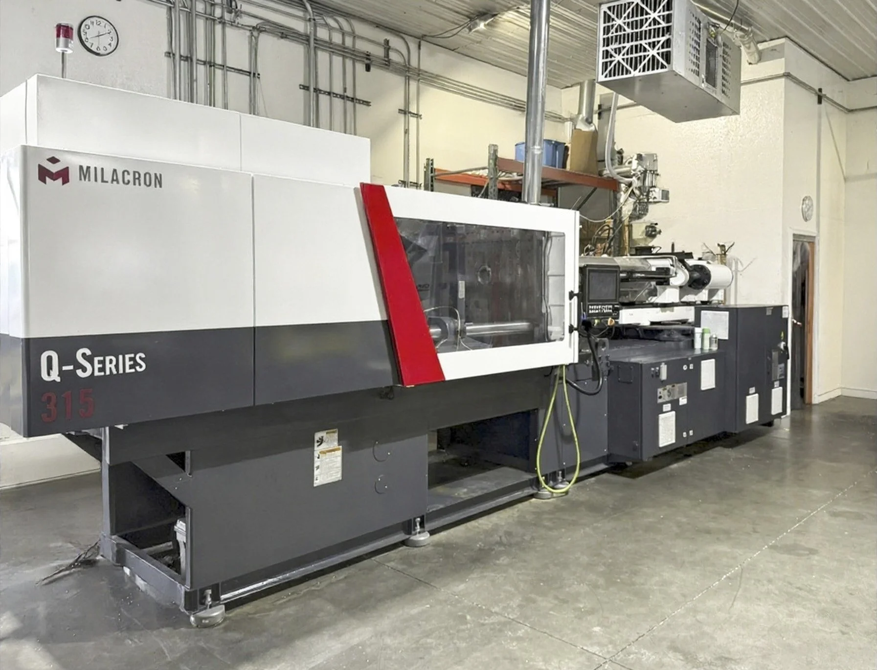 Industrial equipment machine in a factory setting, labeled 'Milacron Q-Series 315', with control panels, wiring, and a transparent cover on part of it.