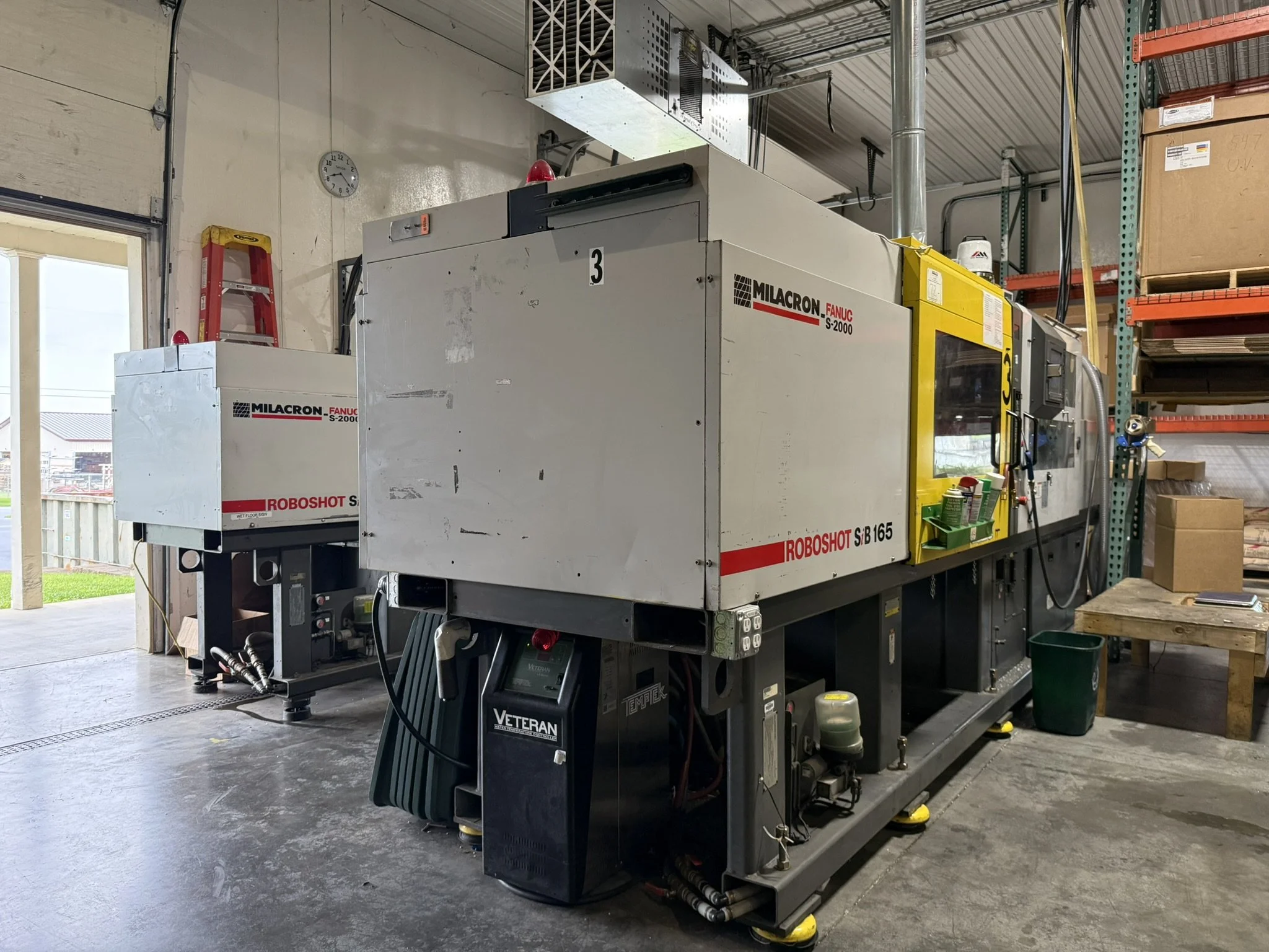 Large industrial machine named Milacron Fanuc S-2000 in a warehouse or workshop with metal shelving filled with boxes and supplies. The machine is white with yellow accents and has a digital control panel. There are ladder and tools nearby, and a clo