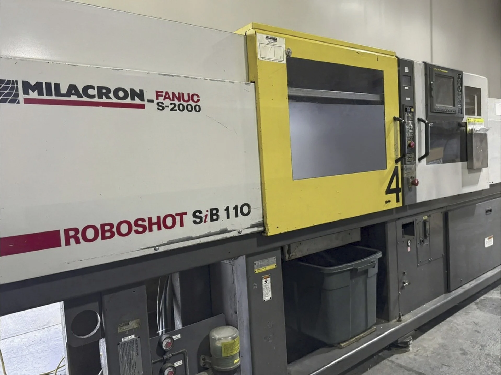 Industrial machine equipment with labels indicating 'Milacron FANUC S-2000' and 'ROBOTSHOT SiB 110'. The machine has a yellow and gray exterior and control panels on the side, situated in a factory or industrial setting.
