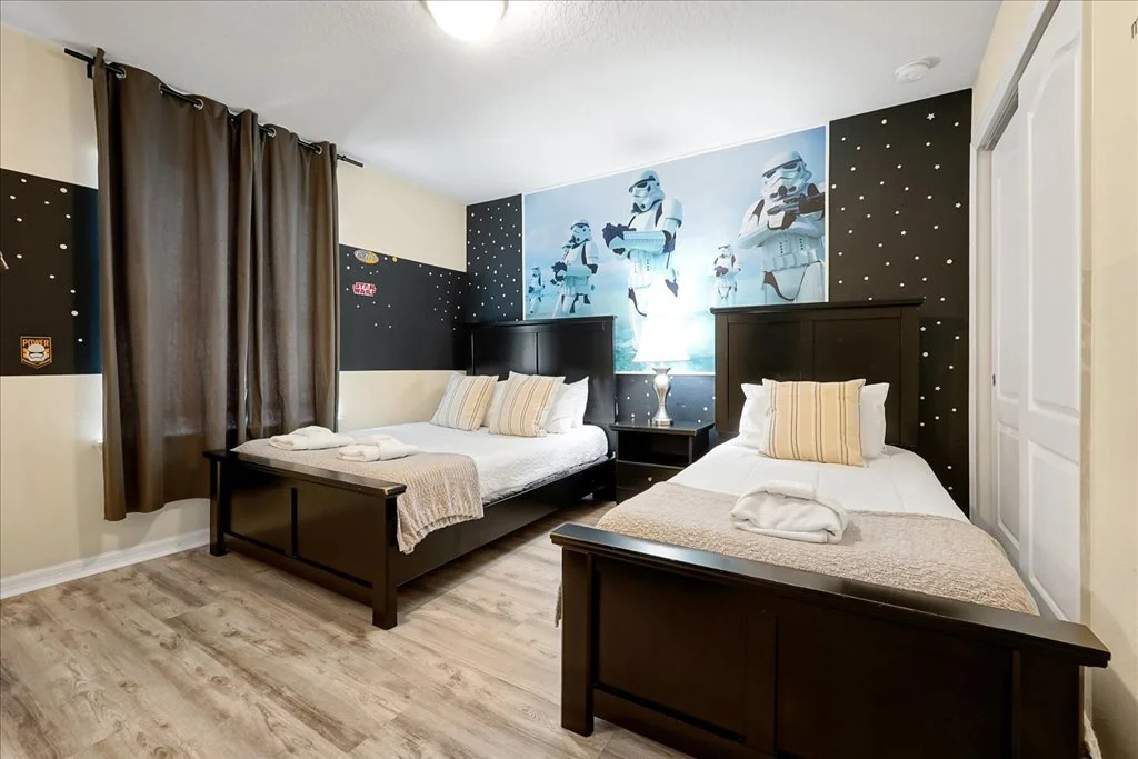 Room with two beds, Star Wars Stormtrooper-themed wall art, black and white decor, wooden flooring, brown curtains, and a nightstand with a lamp.