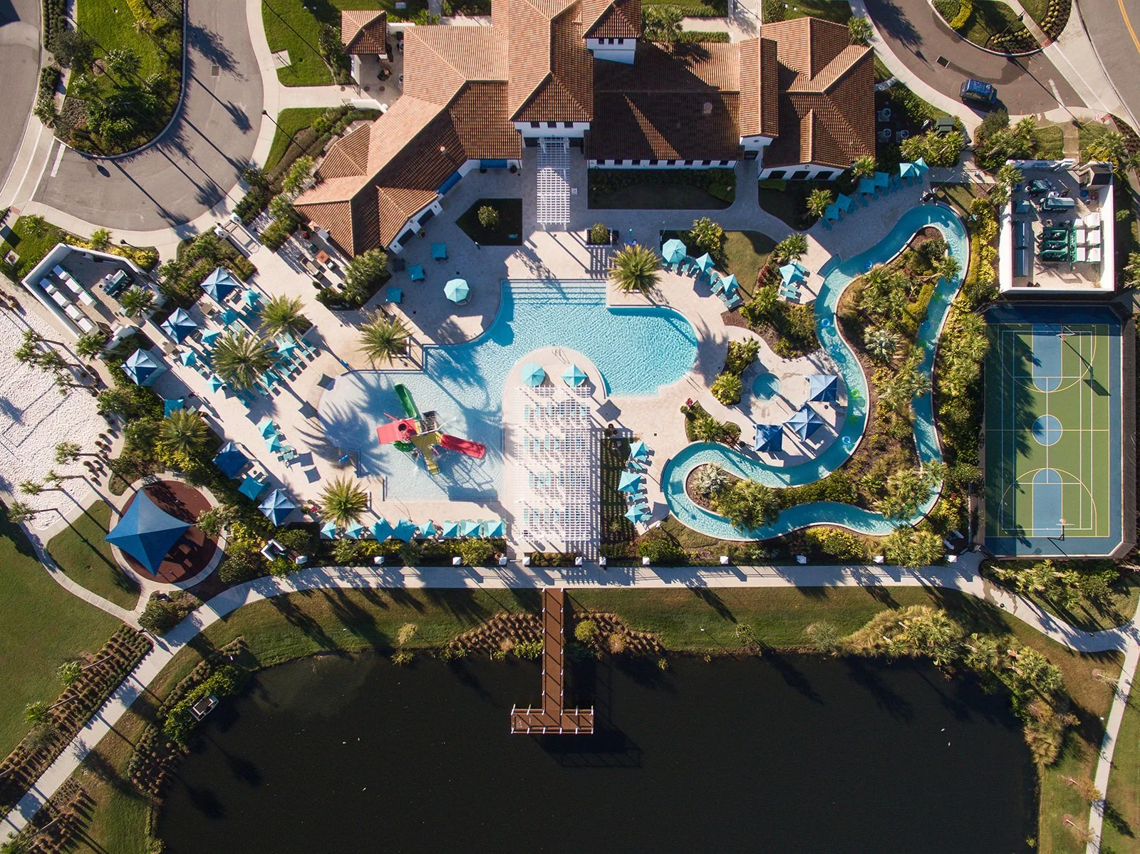 Aerial view of a large residential community pool area with a lazy river, multiple lounge chairs with umbrellas, a playground, a sports court, and surrounding trees and pathways.