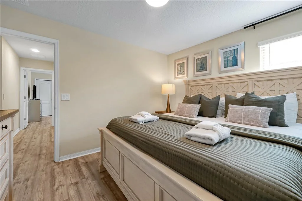 A neatly made bed with multiple pillows in a bedroom, with a nightstand and lamp, artwork on the wall, and a view into an adjacent room.