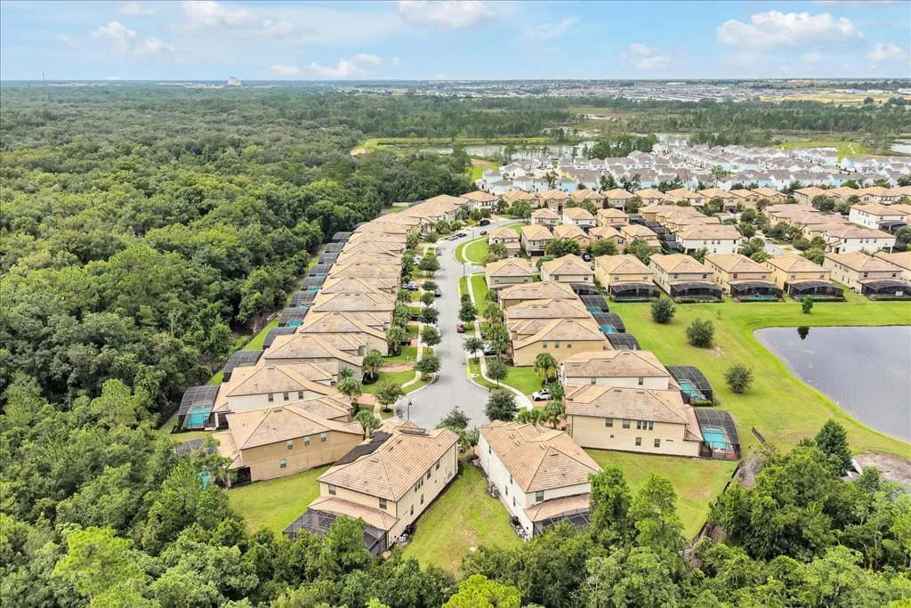 Aerial view of a suburb with rows of beige houses, green lawns, and a pond, surrounded by wooded area.