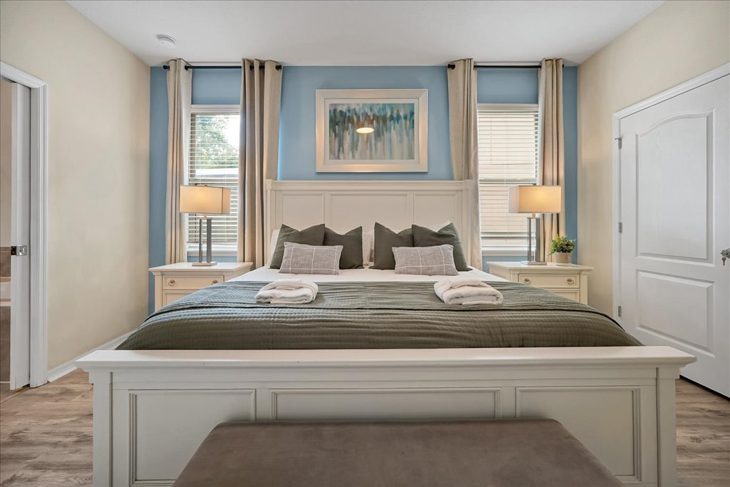 A neatly made bed with a white wooden frame, gray bedding, and multiple gray and beige pillows in a bright, well-lit bedroom. Two nightstands with lamps and curtains are positioned on either side of the bed, with a painting hanging above the headboar