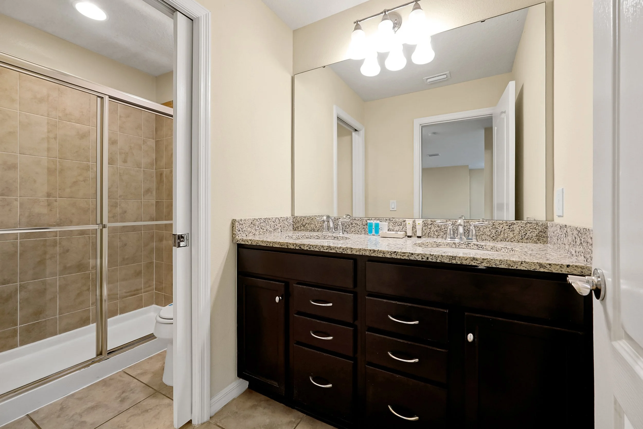 Bathroom with a granite countertop vanity, dark wood cabinets, large mirror, three-light fixture, and a separate shower with glass door and beige tile walls.