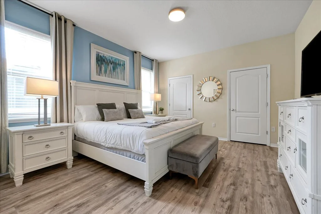 A bedroom with a large white bed, nightstands with lamps, an abstract painting above the headboard, a circular mirror on the wall, a gray bench at the foot of the bed, two windows with beige curtains, and a dresser with a television.