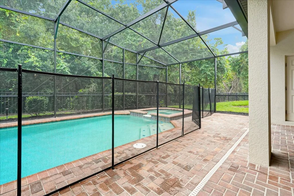Enclosed backyard swimming pool with safety netting, brick patio, and view of trees and blue sky.