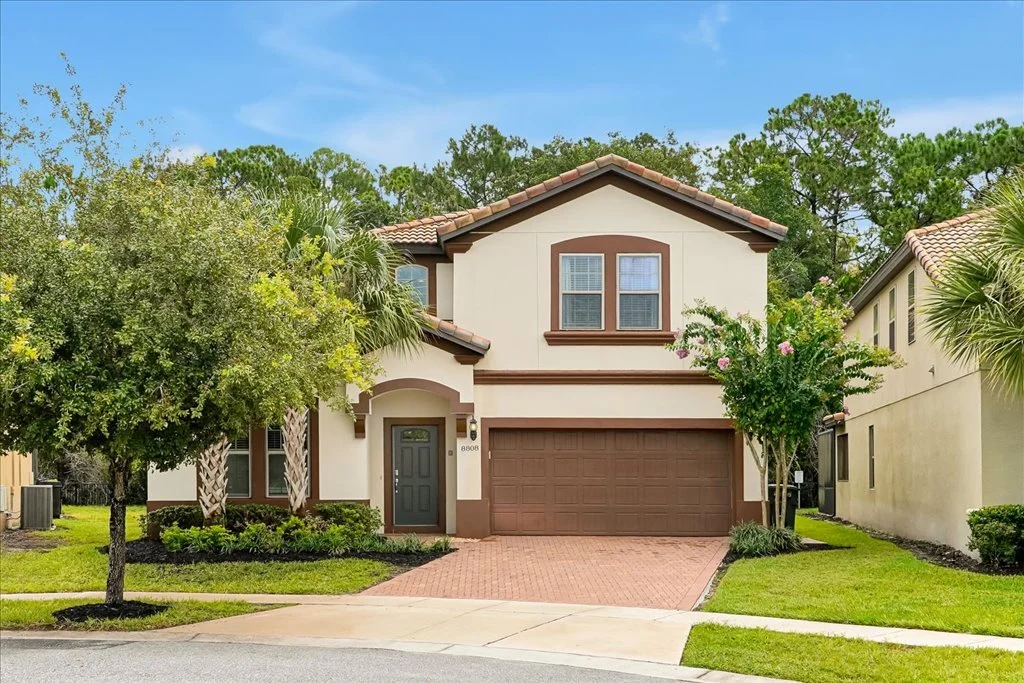 A two-story suburban house with a beige and brown exterior, a two-car garage, and a small front yard with trees and bushes under a blue sky.