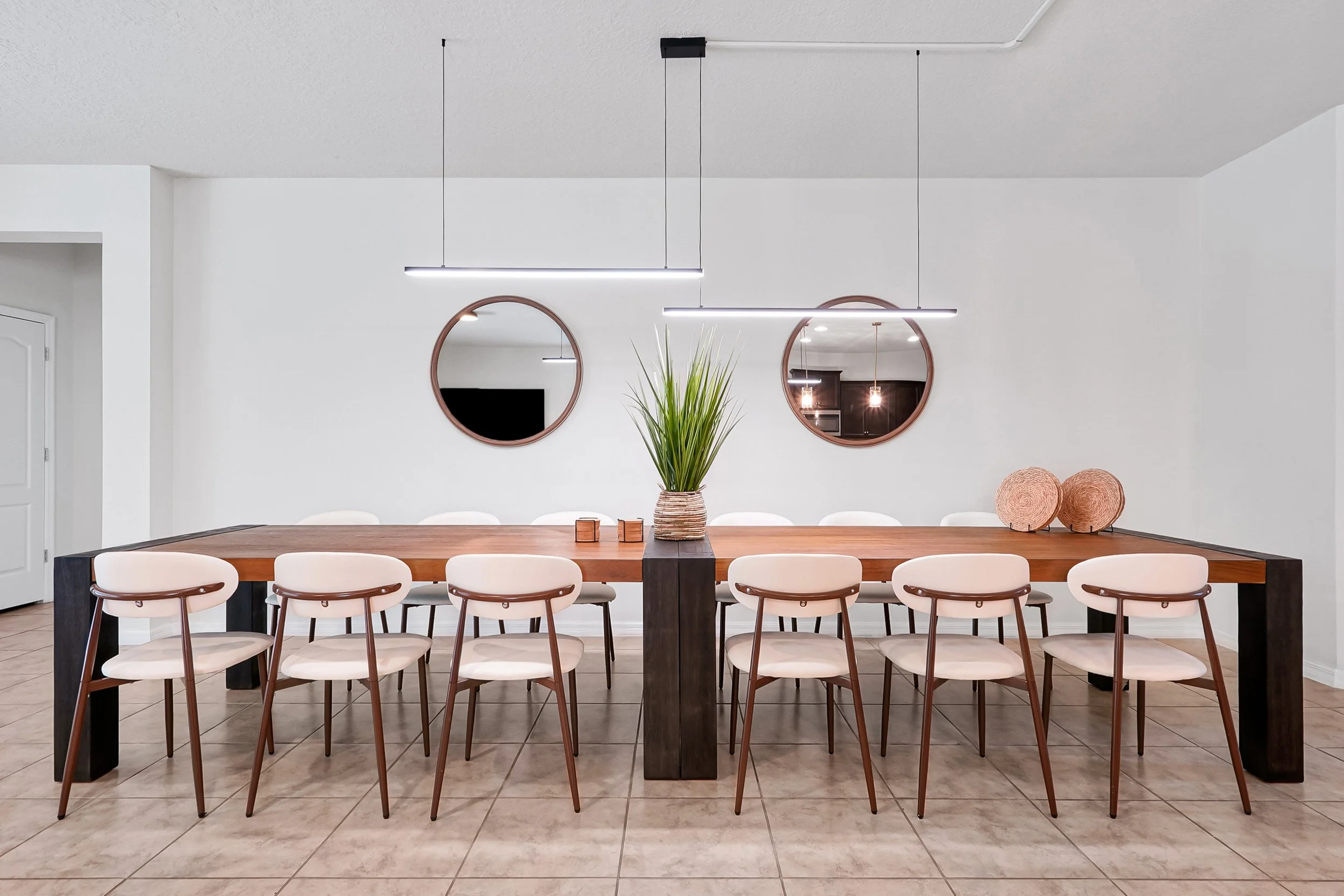 A modern dining room with a wood dining table surrounded by white chairs with brown accents. There are two round mirrors on the white wall behind the table, a tall green plant in the center, and contemporary black pendant lights hanging from the ceil
