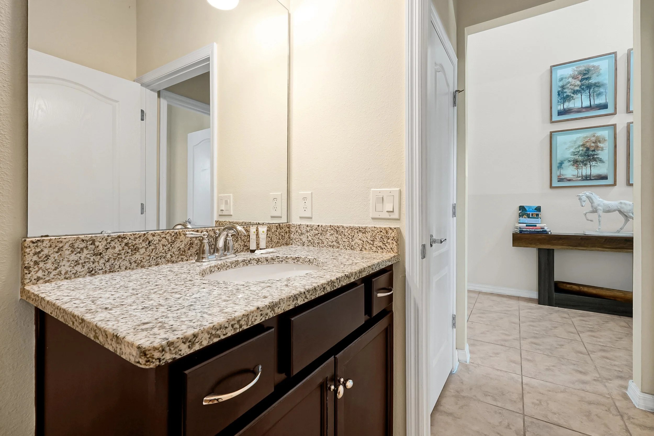 A bathroom vanity with a granite countertop, sink, and cabinet, reflected in a large mirror. In the background, part of another room is visible with framed pictures of trees and a decorative white horse sculpture on a table.