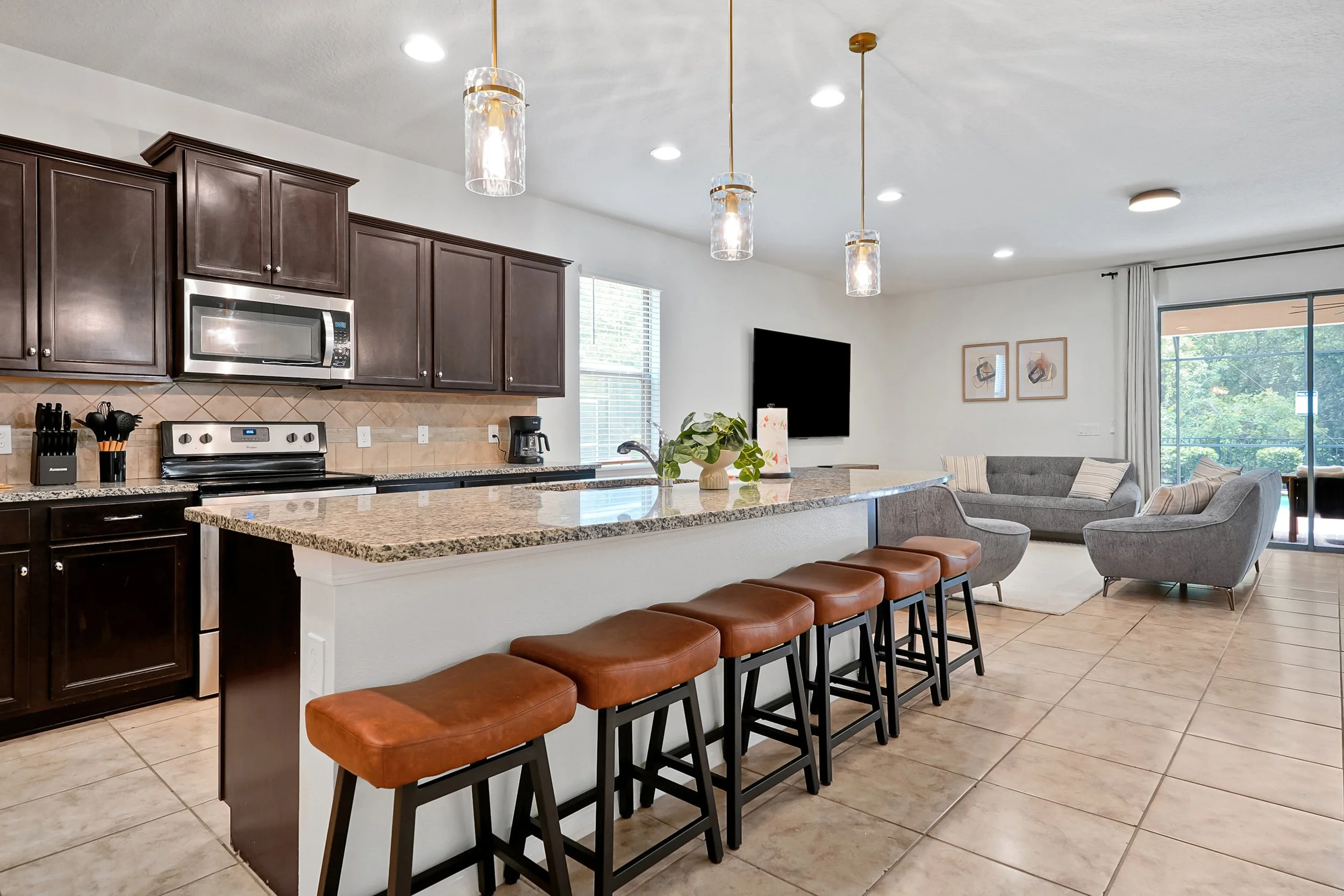 A modern kitchen and living room with dark wood cabinets, a stainless steel microwave, an electric stove, granite countertops, a coffee maker, and a beige tile floor. There are three pendant lights hanging above the kitchen island, with four leather 