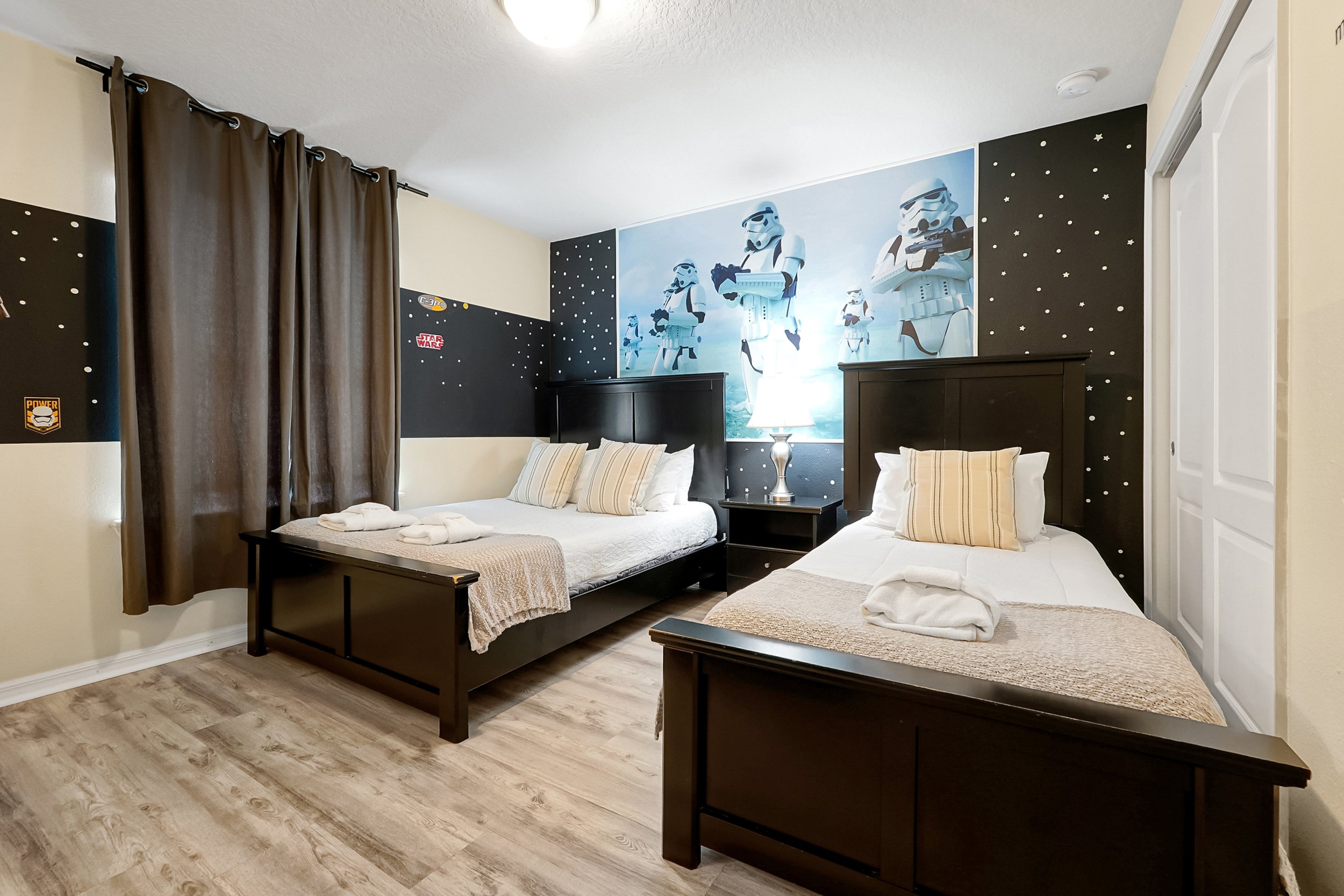 A bedroom with two beds, themed around Star Wars, featuring a large Stormtrooper mural on the wall. The room has black and white decorated walls, wooden furniture, and two bedside tables with lamps.