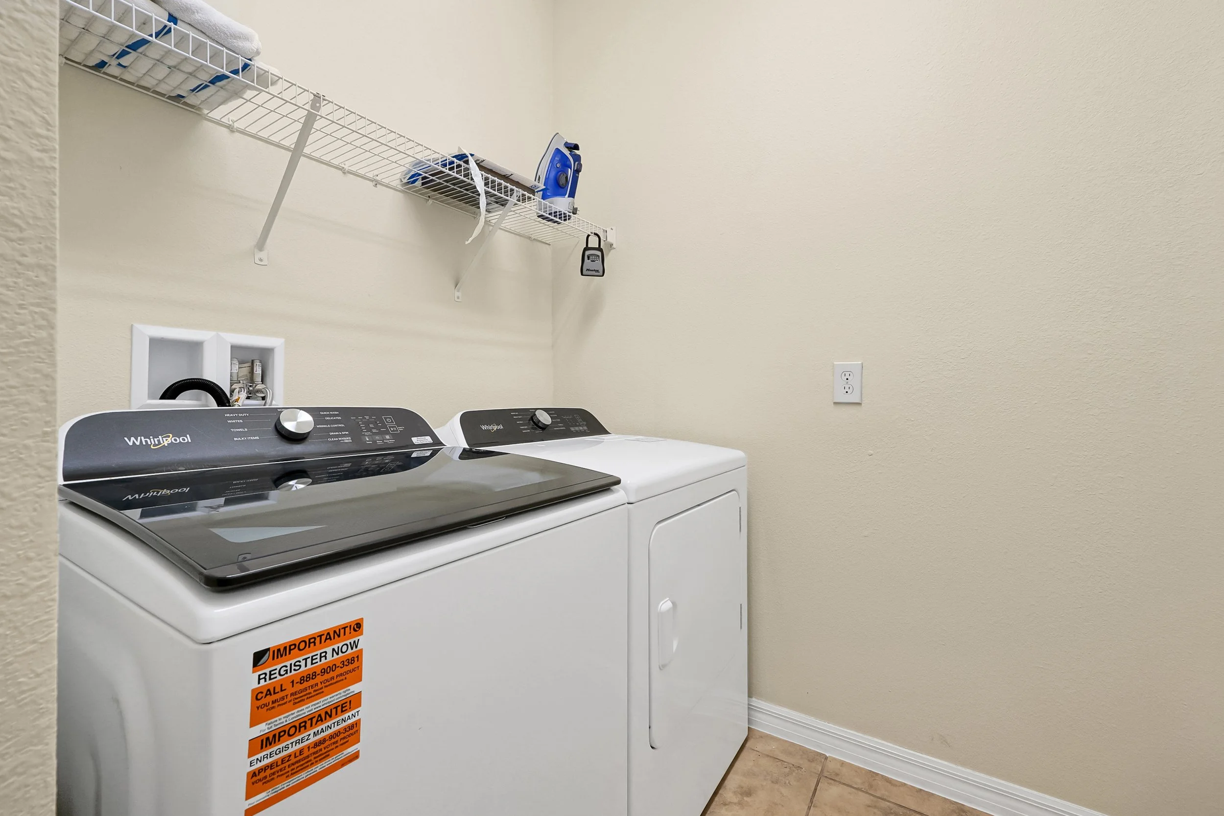 Laundry room with a washing machine and dryer, a wire shelf with a blue iron, a box, and a padlock, and a wall outlet.