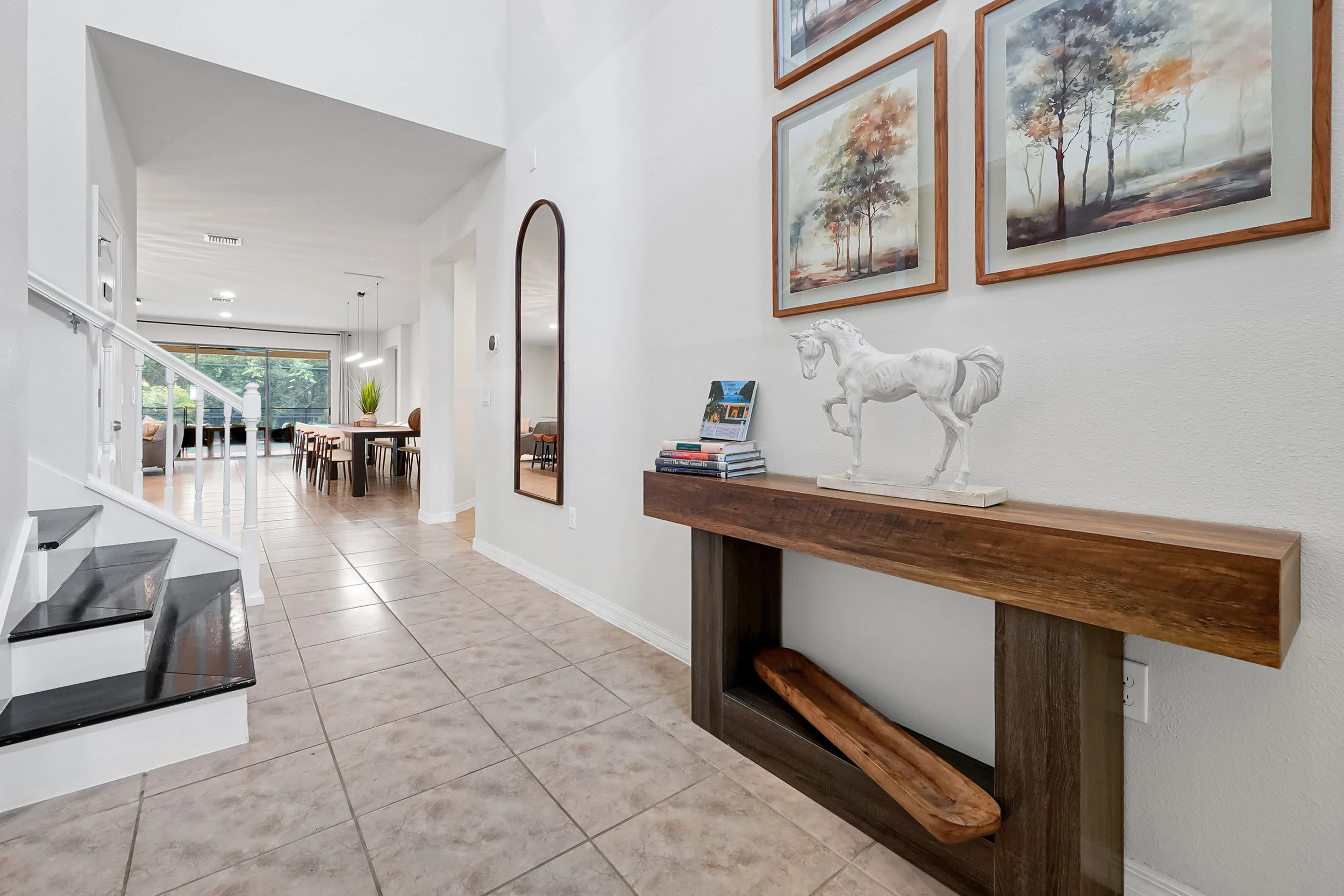 Interior of a house with a tiled floor, white walls, a staircase with black steps, and a hallway leading to a dining and living area with sliding glass doors. Decor includes framed landscape paintings and a white horse sculpture on a wooden console t