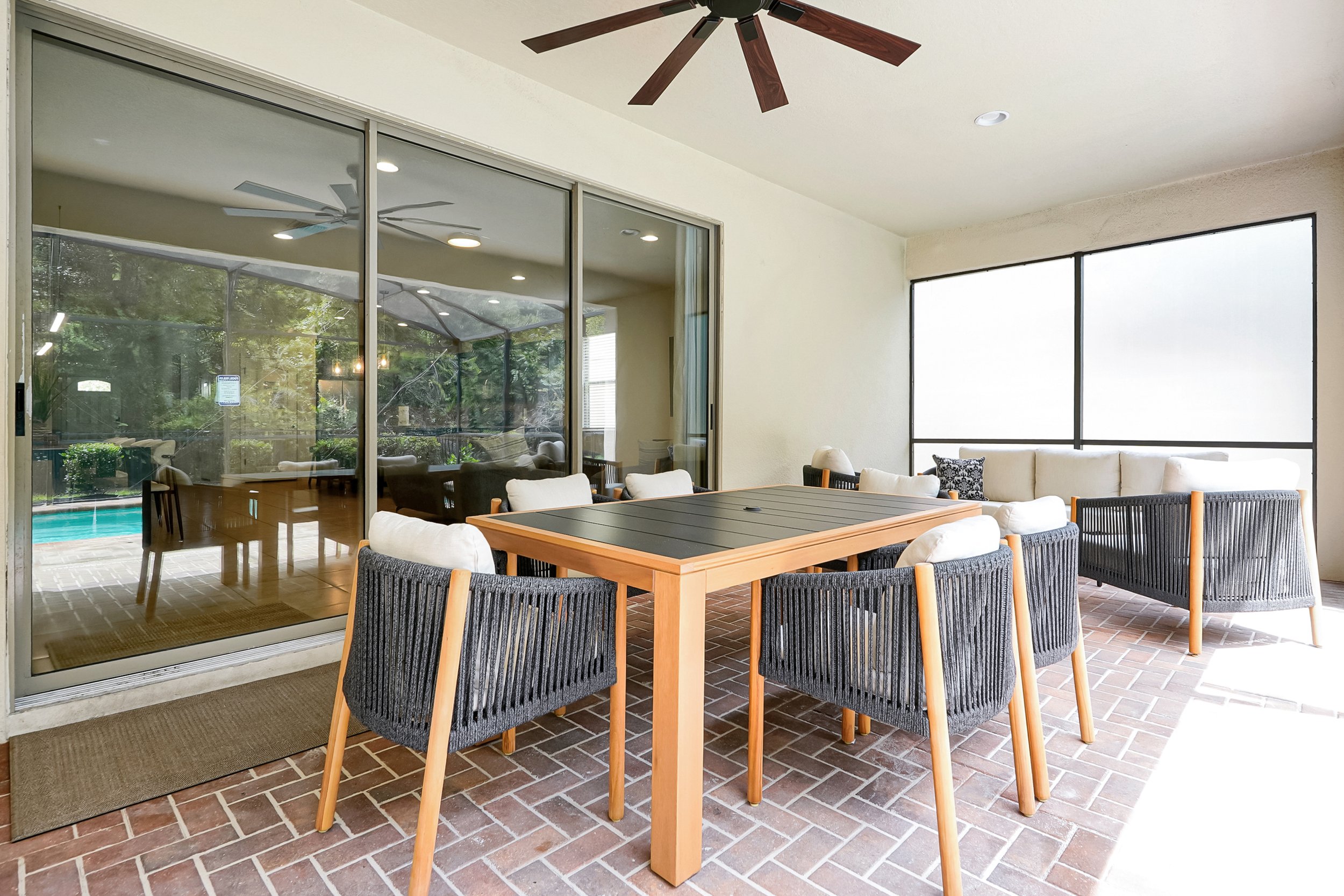 An outdoor patio area with a wooden dining table surrounded by black woven chairs, a ceiling fan, large sliding glass doors, a brick floor, and a sitting area with a white sectional sofa and decorative pillows.
