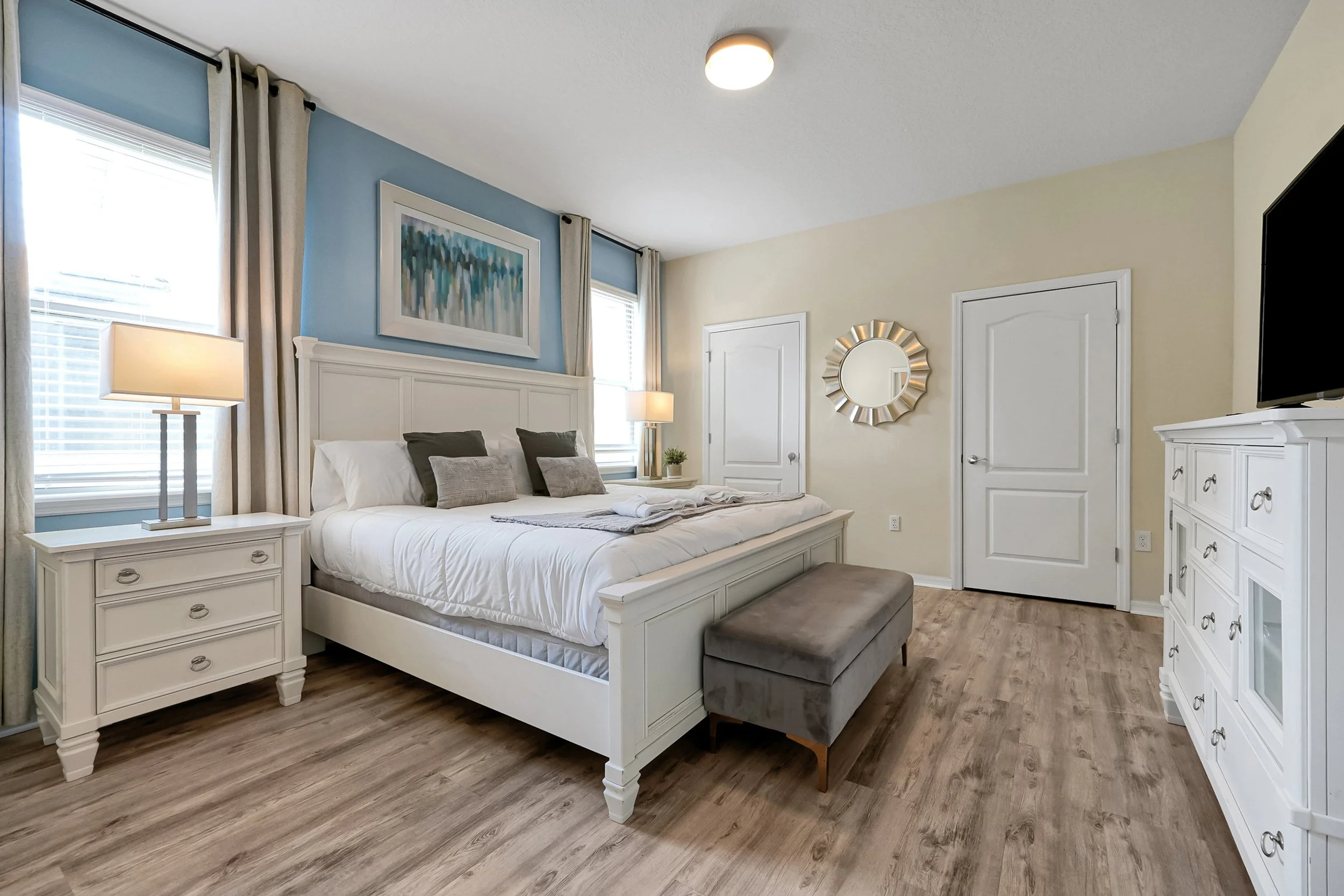A bedroom with a white bed frame and bedding, grey pillows, two nightstands with lamps, a window with beige curtains, a blue and beige wall with a painting, and a white dresser with a TV on top.