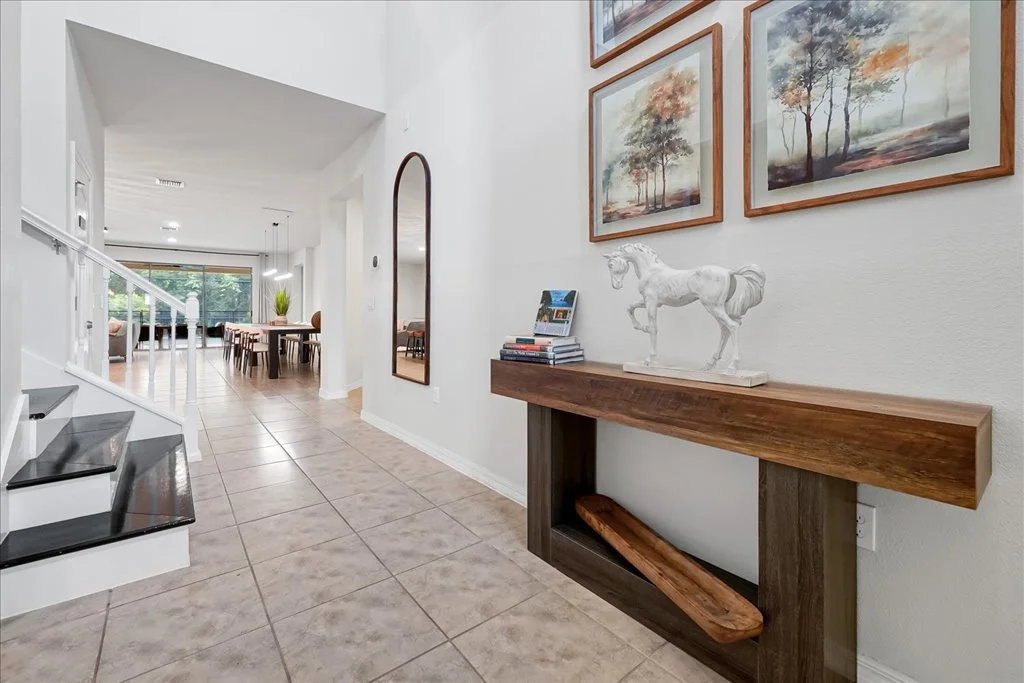 View of a hallway with a staircase on the left, leading to a living area in the background. On the right, a wooden console table features a white horse sculpture, a small stack of books, and a few magazines. Above the table, there are three framed la