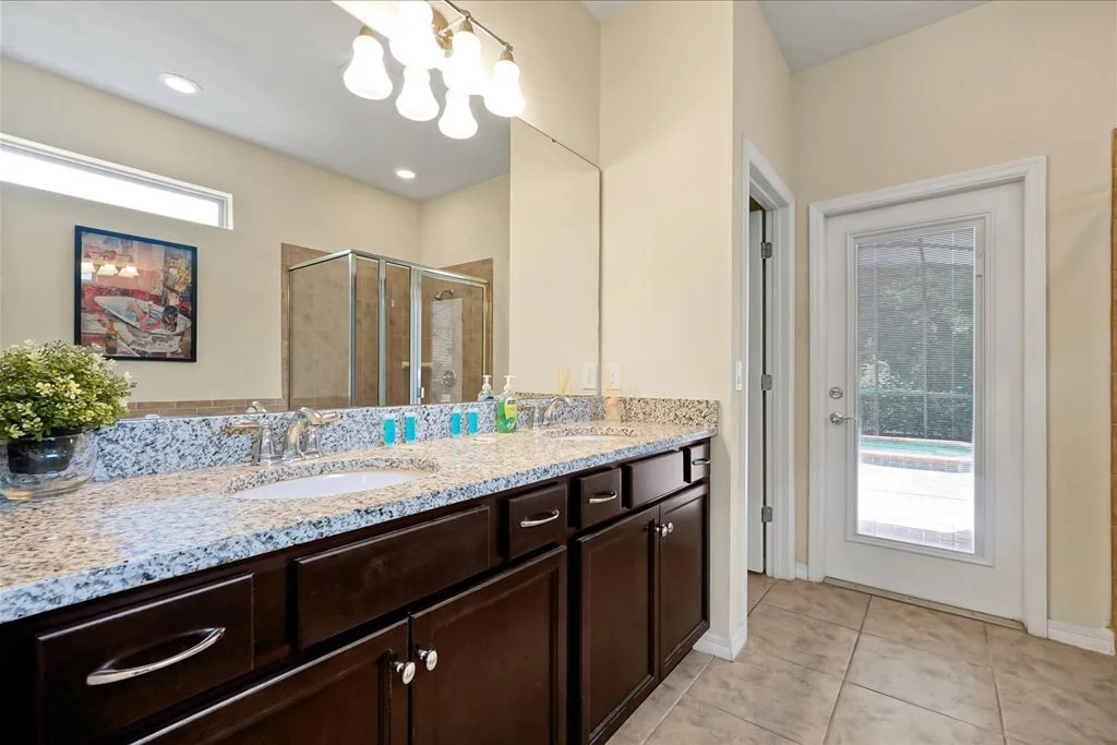 Bathroom with dual sinks, dark wood cabinets, granite countertop, a large mirror, a shower enclosure, a small plant on the counter, and a door with a window leading outside.