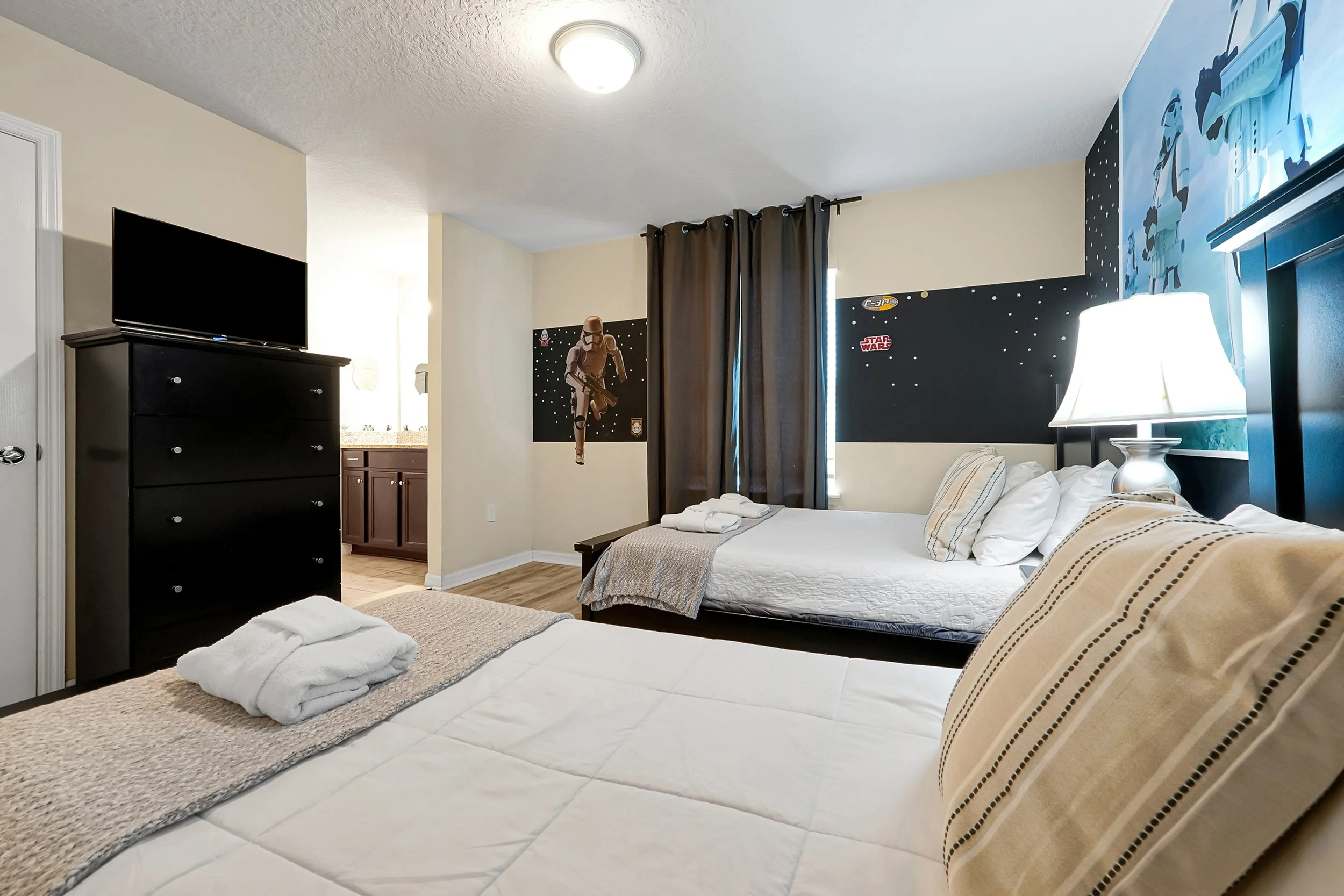 A bedroom with two beds, one in the foreground and one in the background, both with white linens and striped pillows. Towels are folded on a rug beside the beds. A black dresser with a television is against the wall, and there are windows with dark c