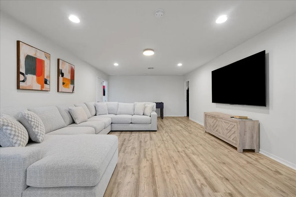 A spacious, modern living room with white walls, a large L-shaped light gray sofa, abstract artwork on the wall, a flat-screen TV mounted on the wall, and wooden flooring.