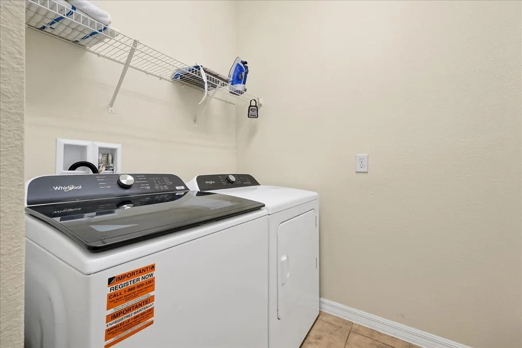 A laundry room with a white washing machine and dryer, a wire shelf above with an iron, and an electrical outlet on the wall.