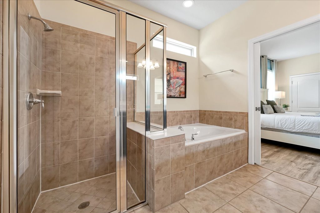 Bathroom with a glass-enclosed shower, a bathtub with tan tiles, a framed artwork, and a doorway leading to a bedroom with a white bed and wooden floor.