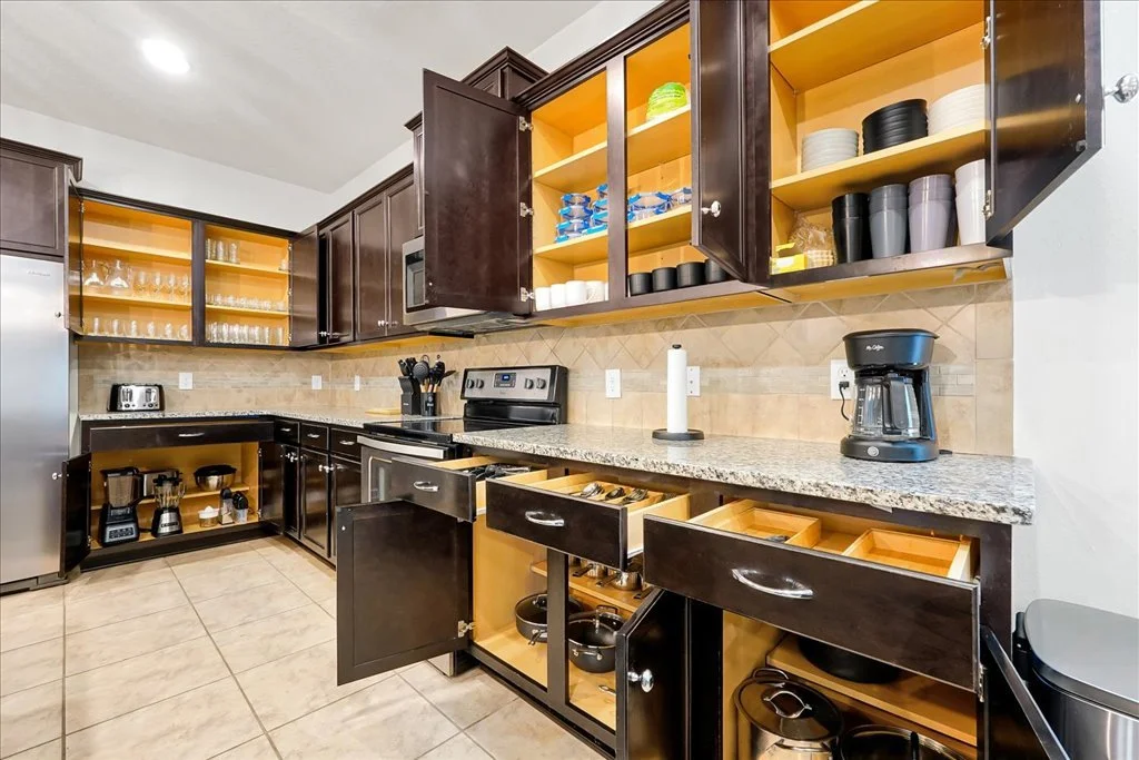 Kitchen cabinets with open and closed drawers, granite countertop, and kitchen appliances including coffee makers, microwave, and stove, with dishes and glasses stored in open cabinets.