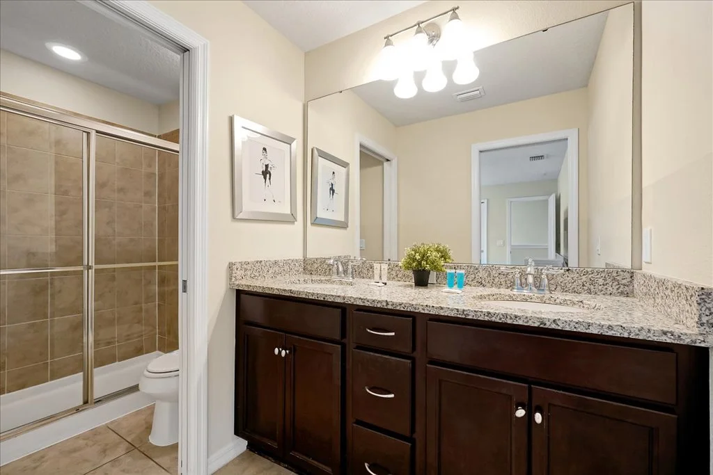 Bathroom with a double sink granite countertop, dark wood cabinets, a large mirror, and a light fixture. There are two framed pictures on the wall. To the left, part of a shower with a sliding glass door and a toilet are visible.